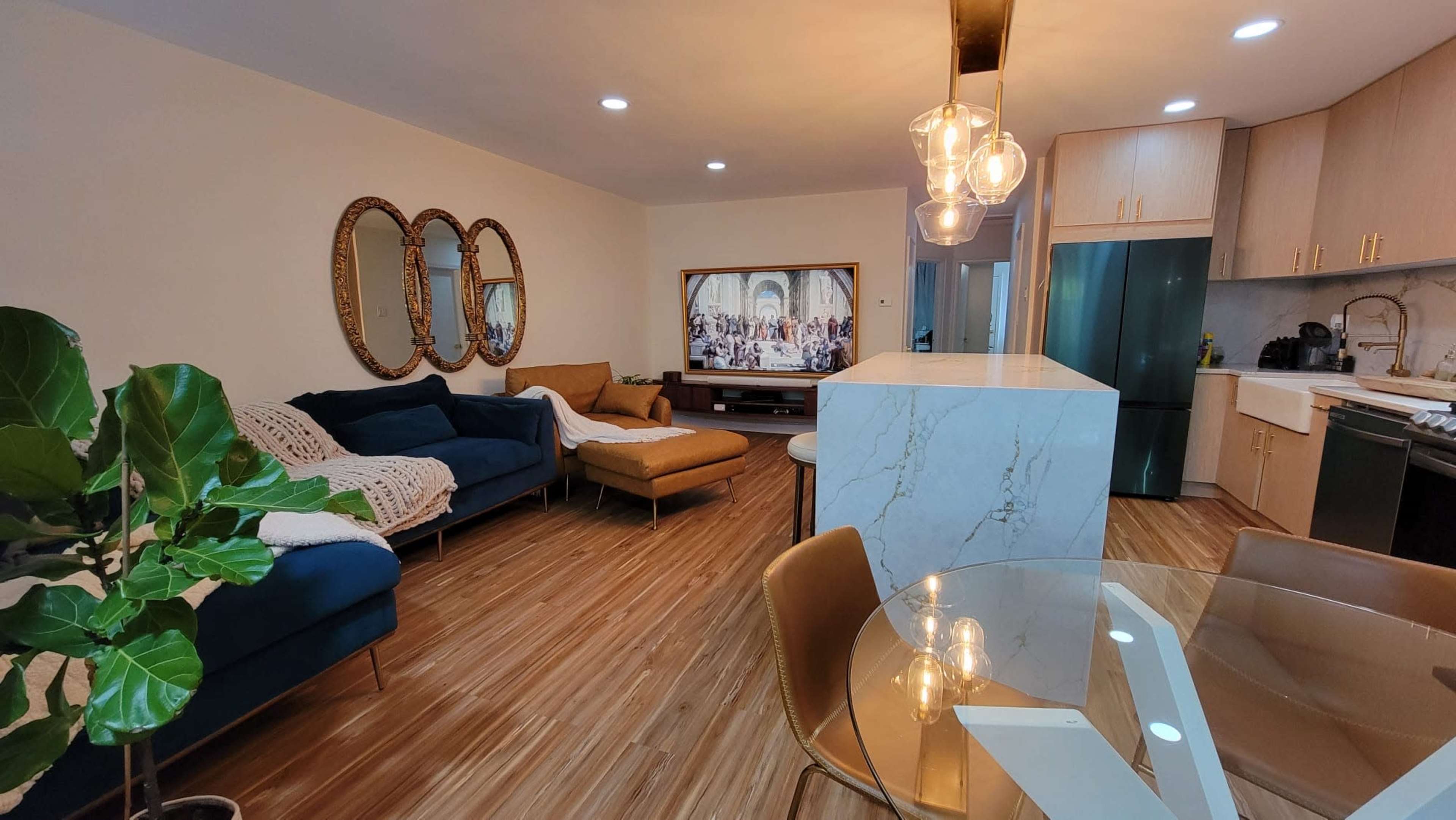 A modern living room and kitchen area with a blue couch, a television mounted on the wall, a marble island, and a dining table.