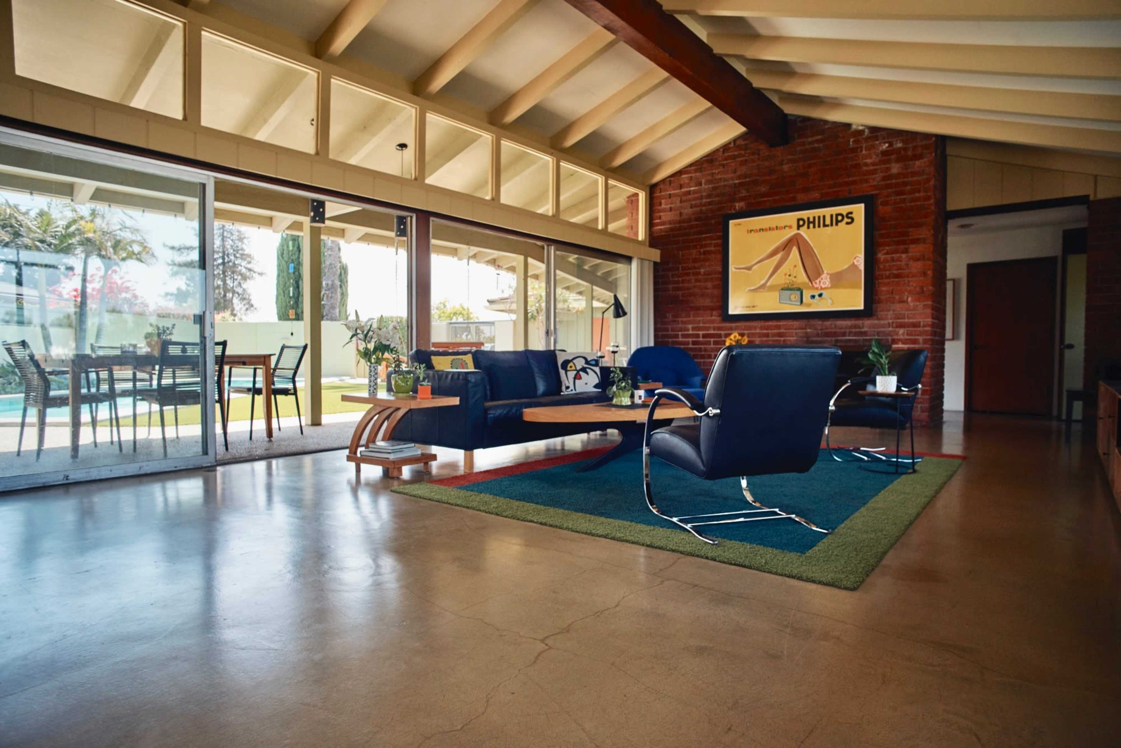 The interior of a modern living room features large windows, a low coffee table, and sleek chairs on a green rug, with a view of an outdoor patio and swimming pool.