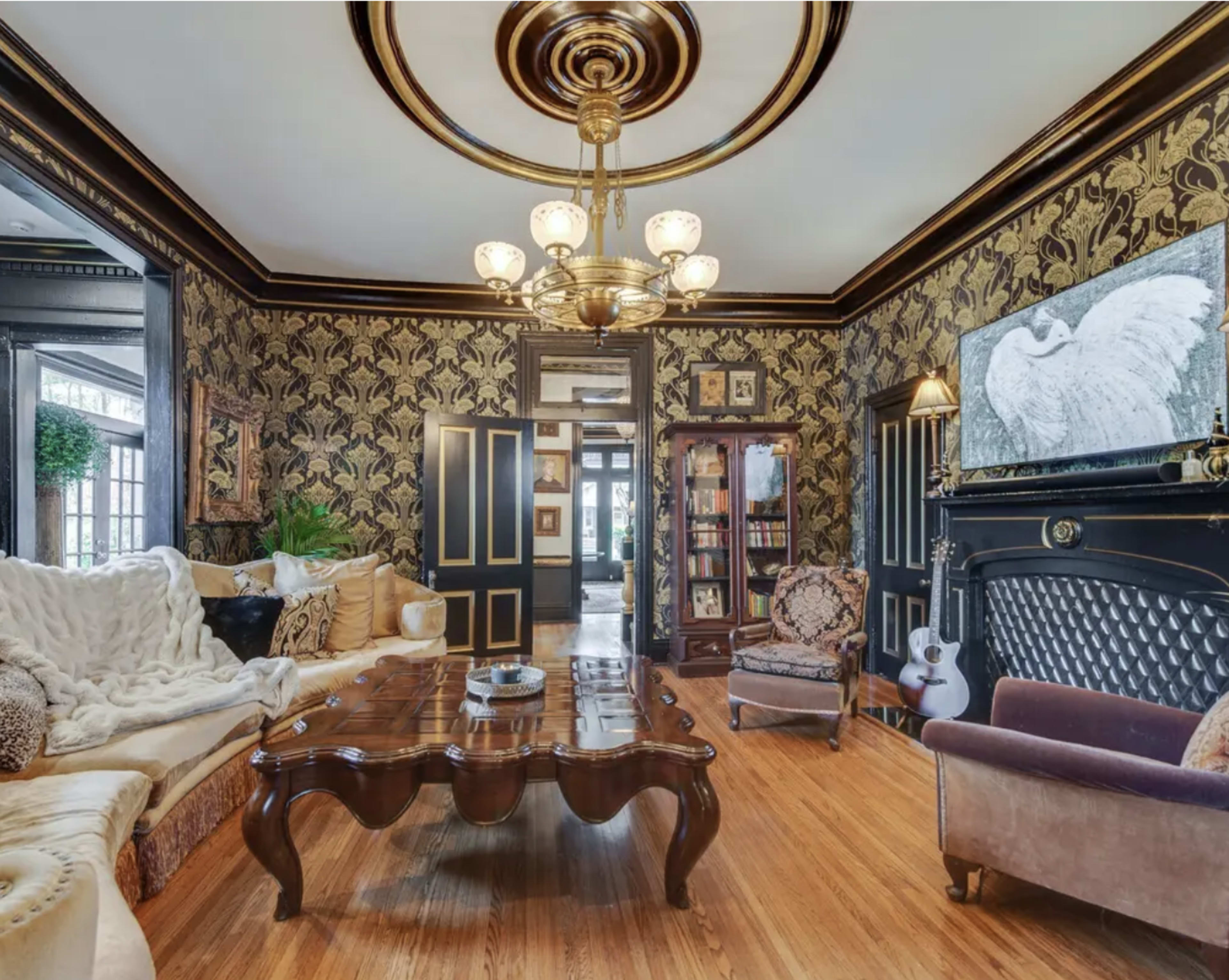 The room features ornate wallpaper, a vintage chandelier, and a mix of seating arrangements around a dark wood coffee table.