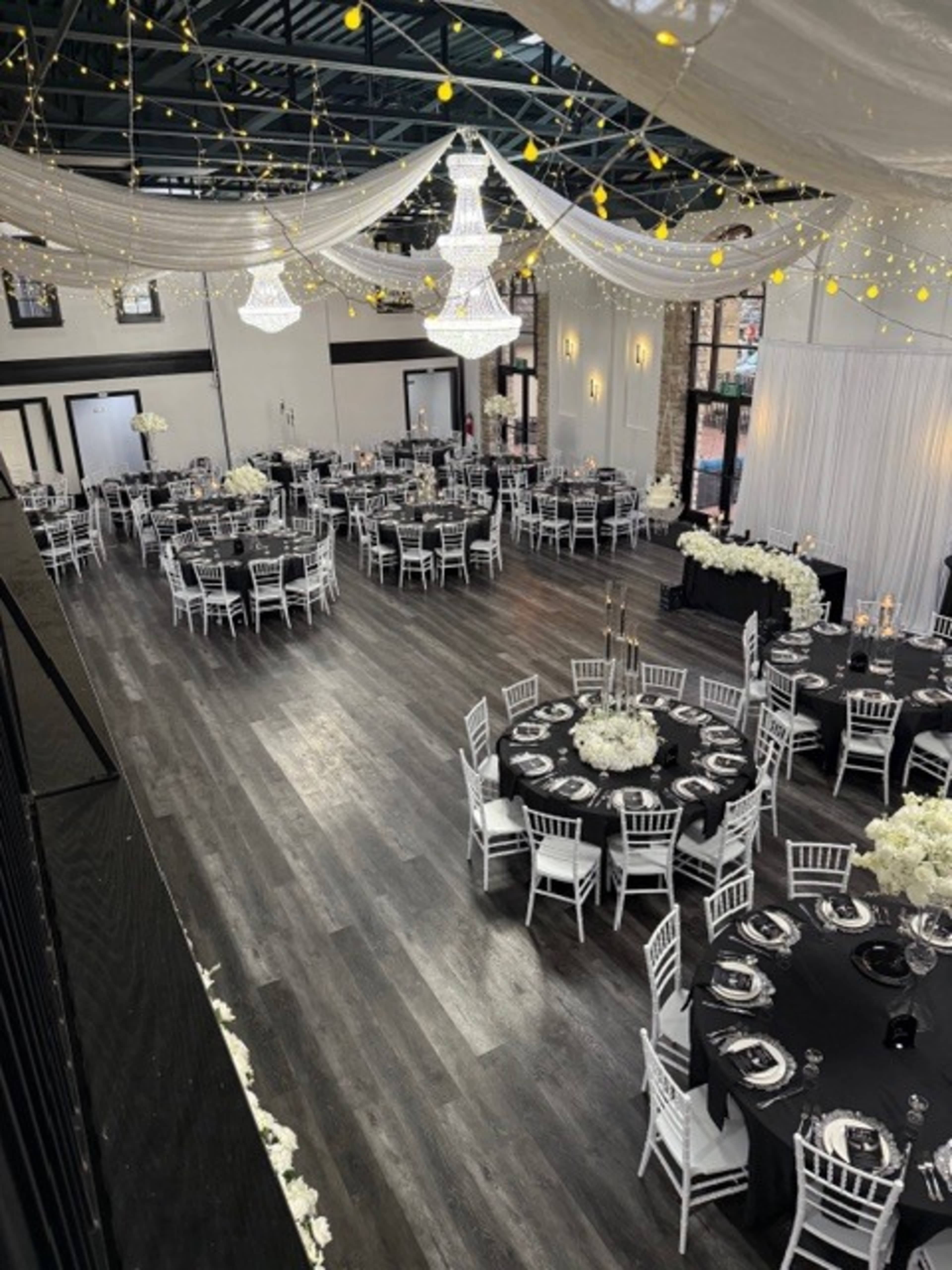 The image shows a spacious event hall set for a formal gathering, featuring elegantly arranged black and white tables adorned with floral centerpieces, surrounded by white chiavari chairs under decorative chandeliers.