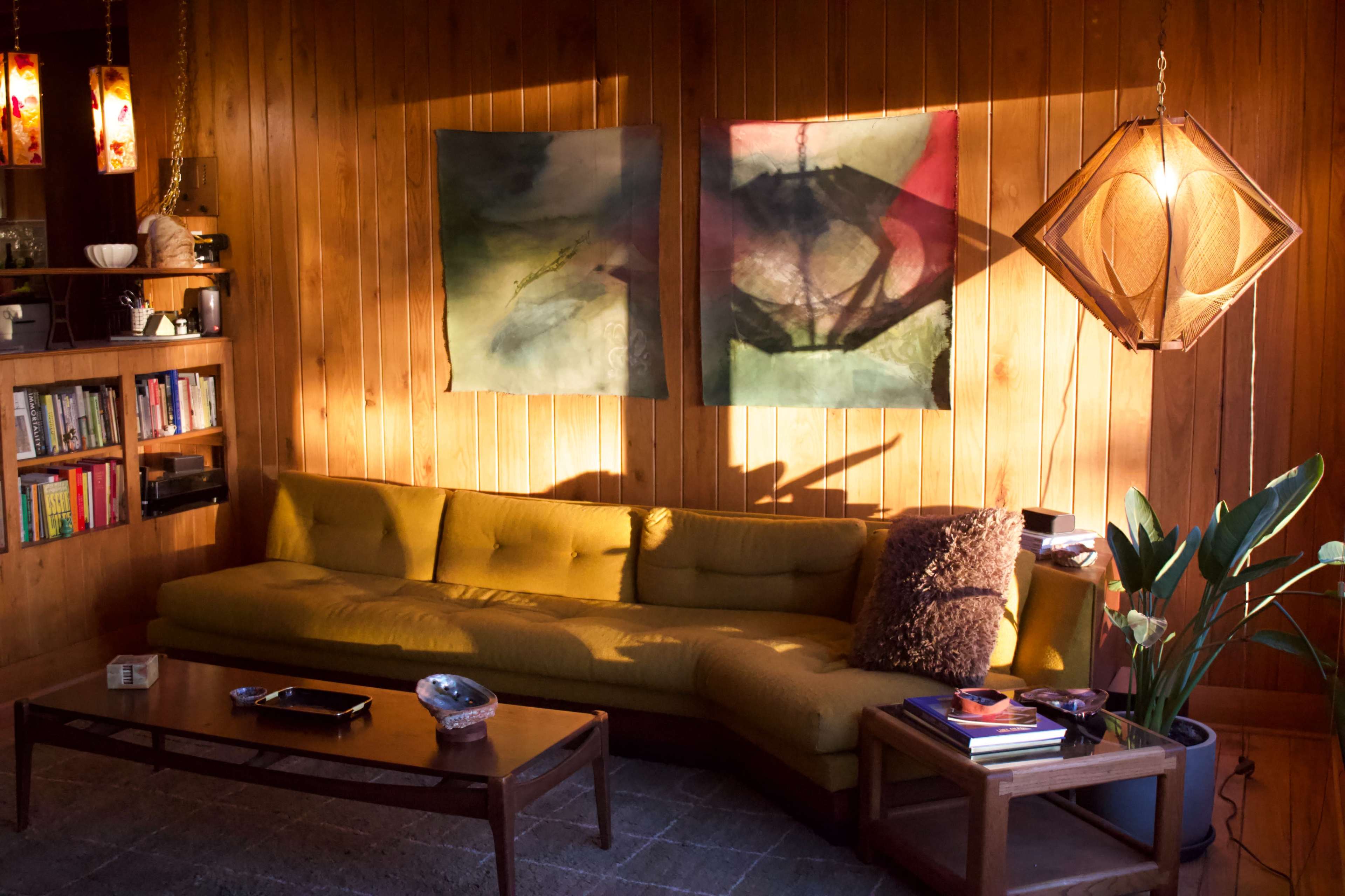 The scene shows a cozy living room with wooden paneling, a yellow couch, two abstract paintings on the wall, and a coffee table with books and decorative items.