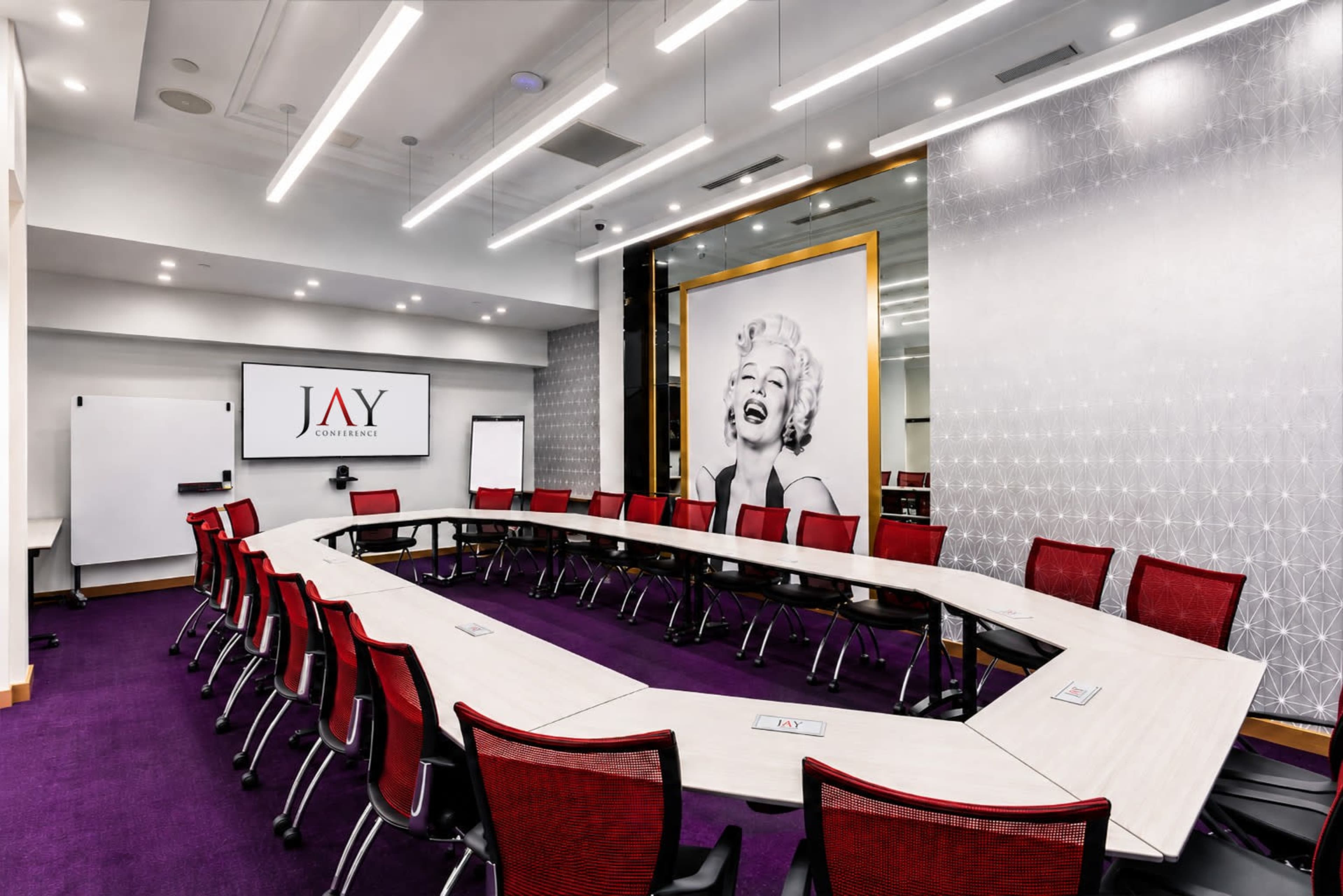 The image shows a modern conference room featuring a long table arranged in a U-shape, surrounded by red chairs, with a large artwork of a woman on the wall and a presentation screen displaying the name "JAY."