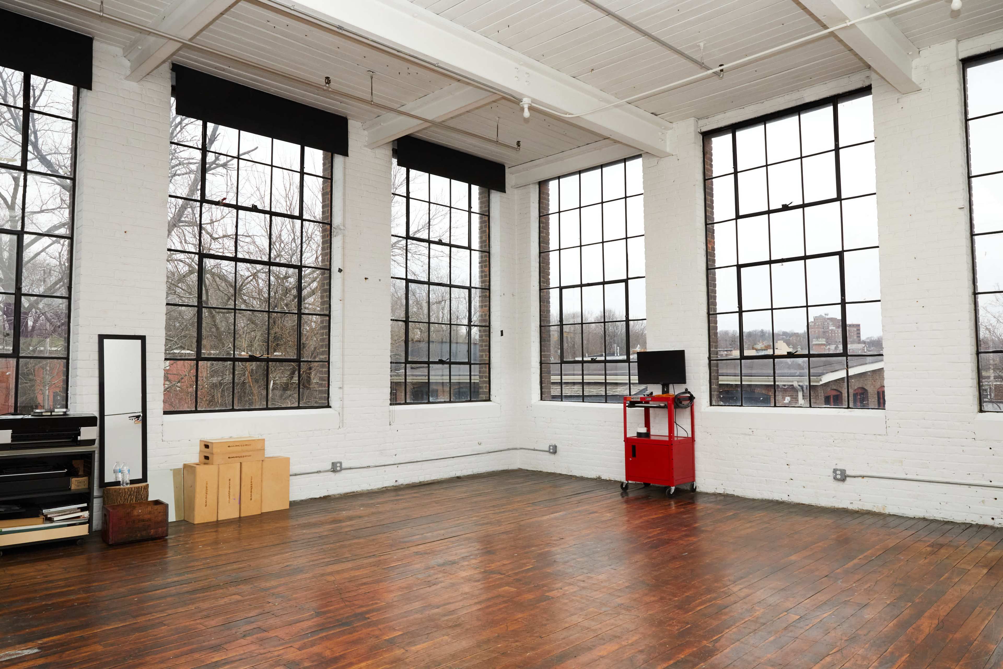 A spacious room with large windows, hardwood floors, and minimal furniture, including a mirror, storage boxes, and a red cart with a television.