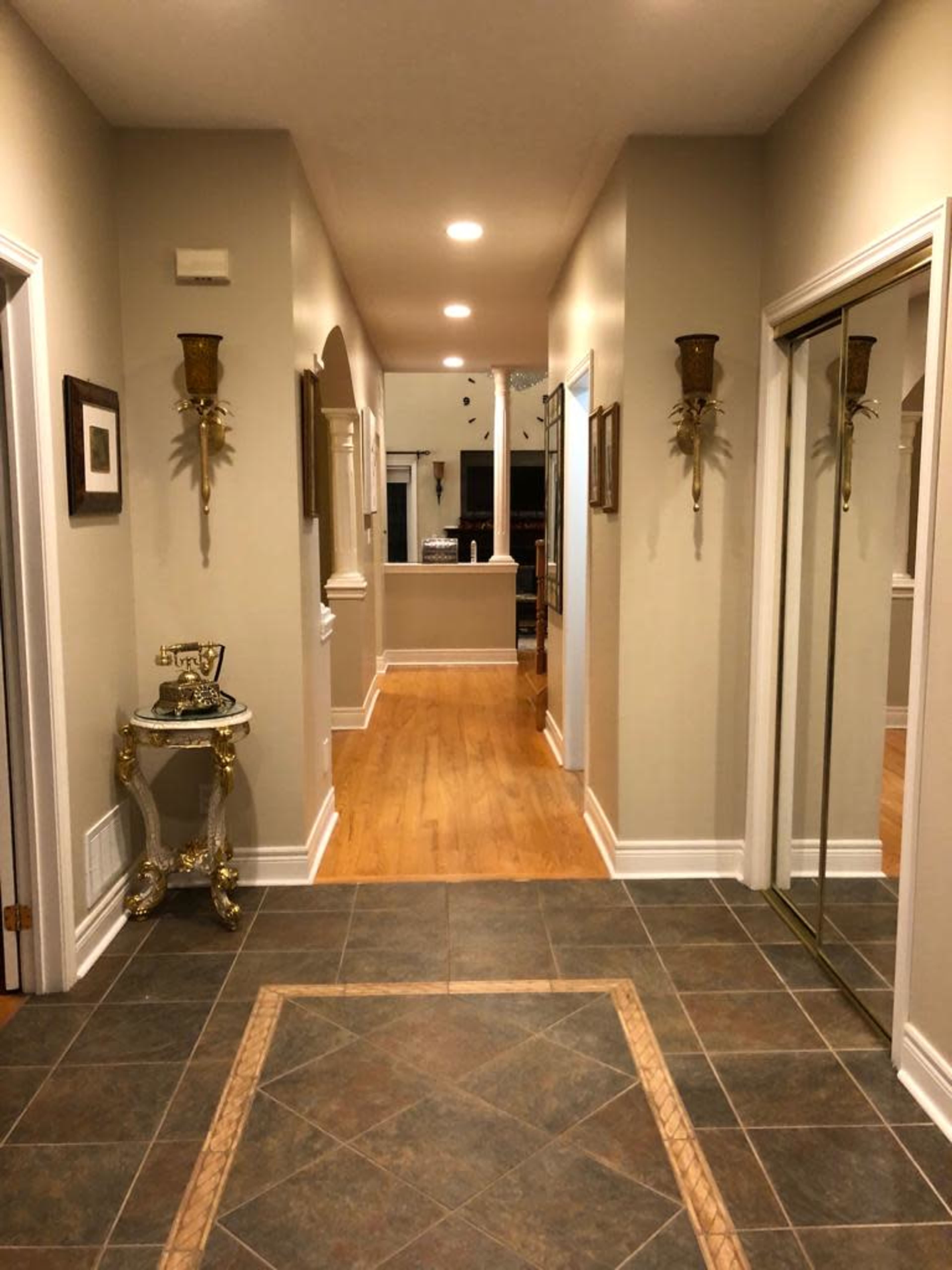 A hallway with tiled flooring, wall sconces, and a mirror leads to a brightly lit area at the end.