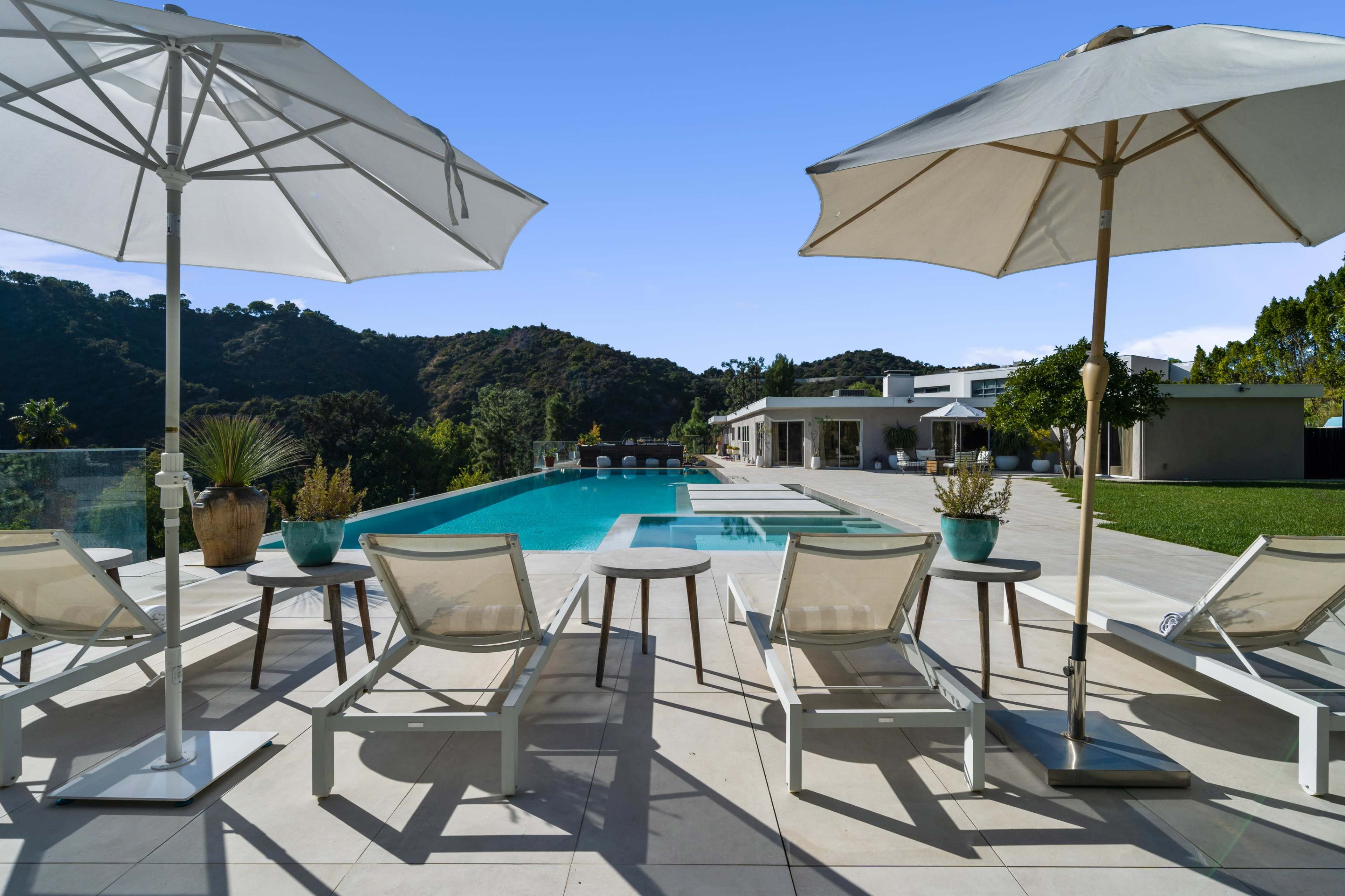 A pool area features white lounge chairs and umbrellas, overlooking a landscaped yard and hills in the background.