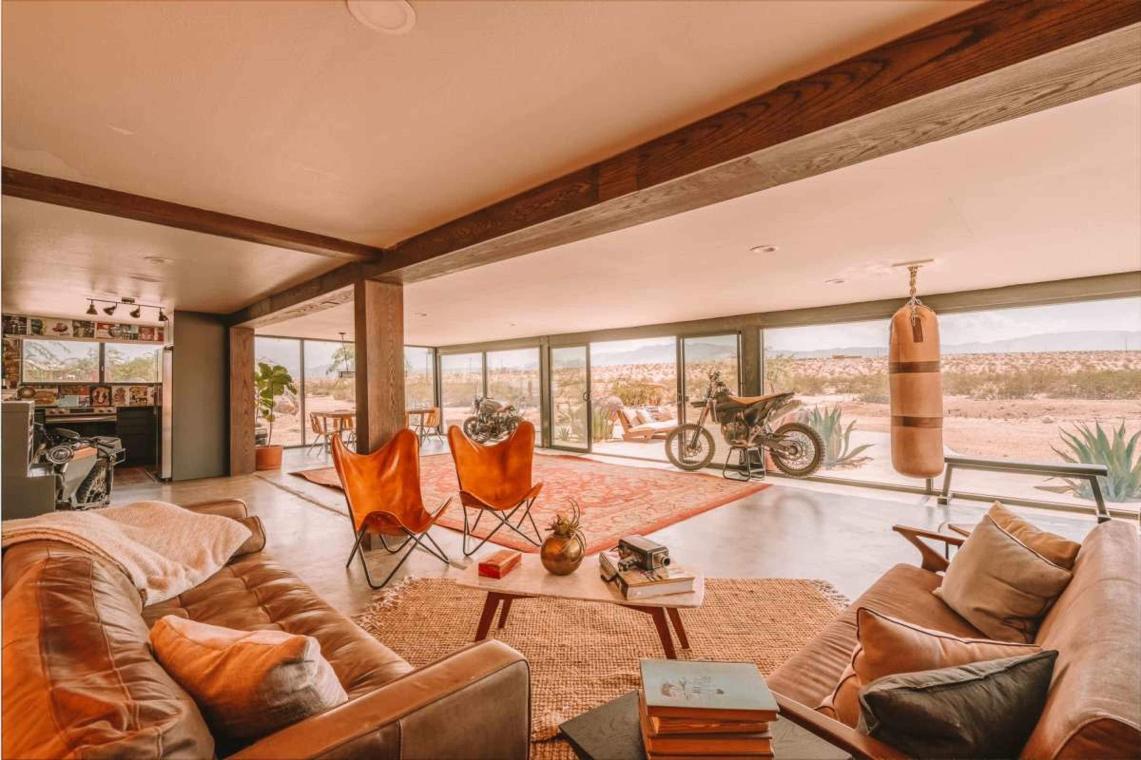 The image showcases a modern living space with large windows overlooking a desert landscape, featuring a leather sofa, two butterfly chairs, and a standing punching bag.