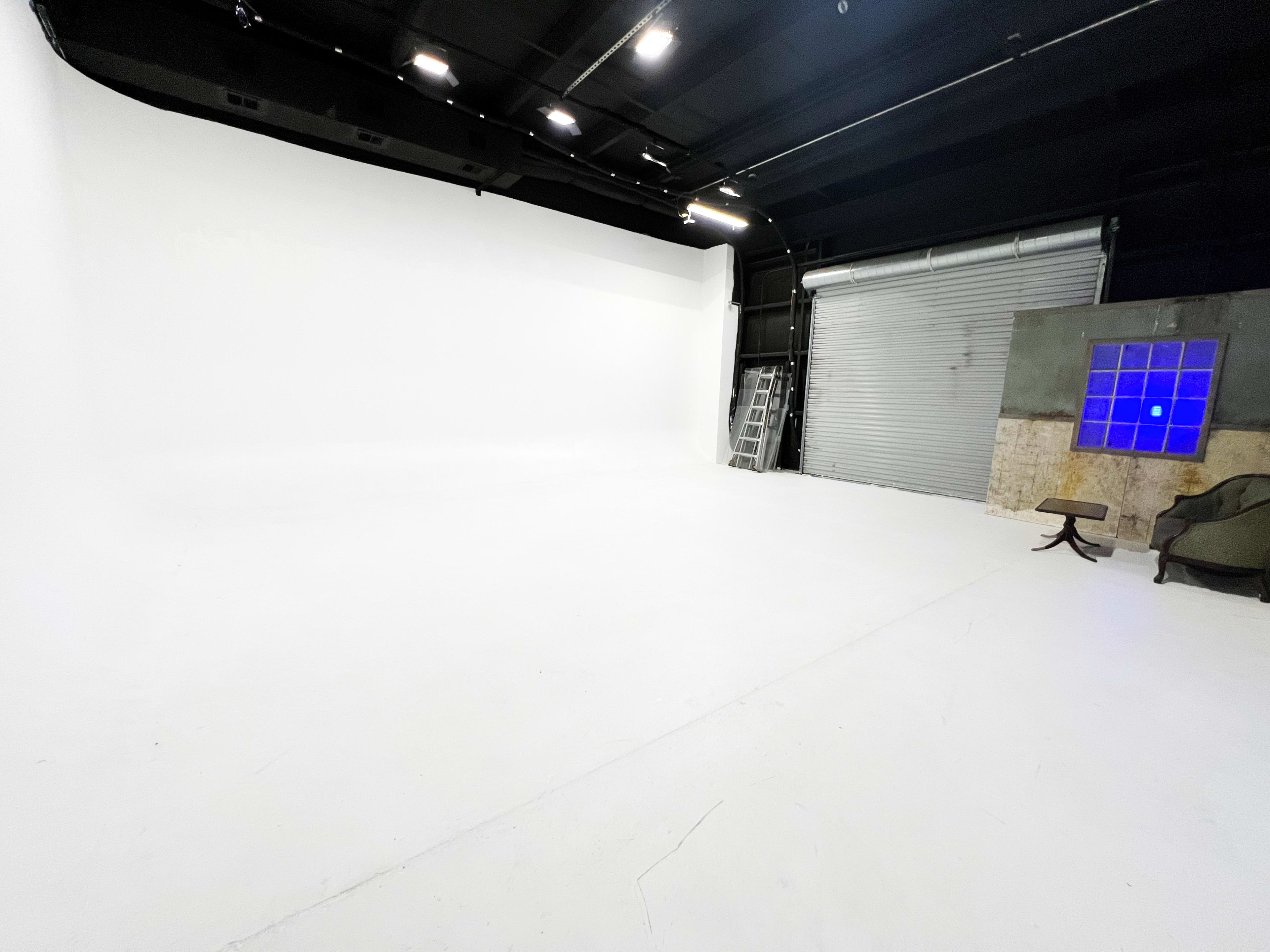 A spacious, empty studio with a white backdrop, metal roll-up door, and a small blue-tinted window.