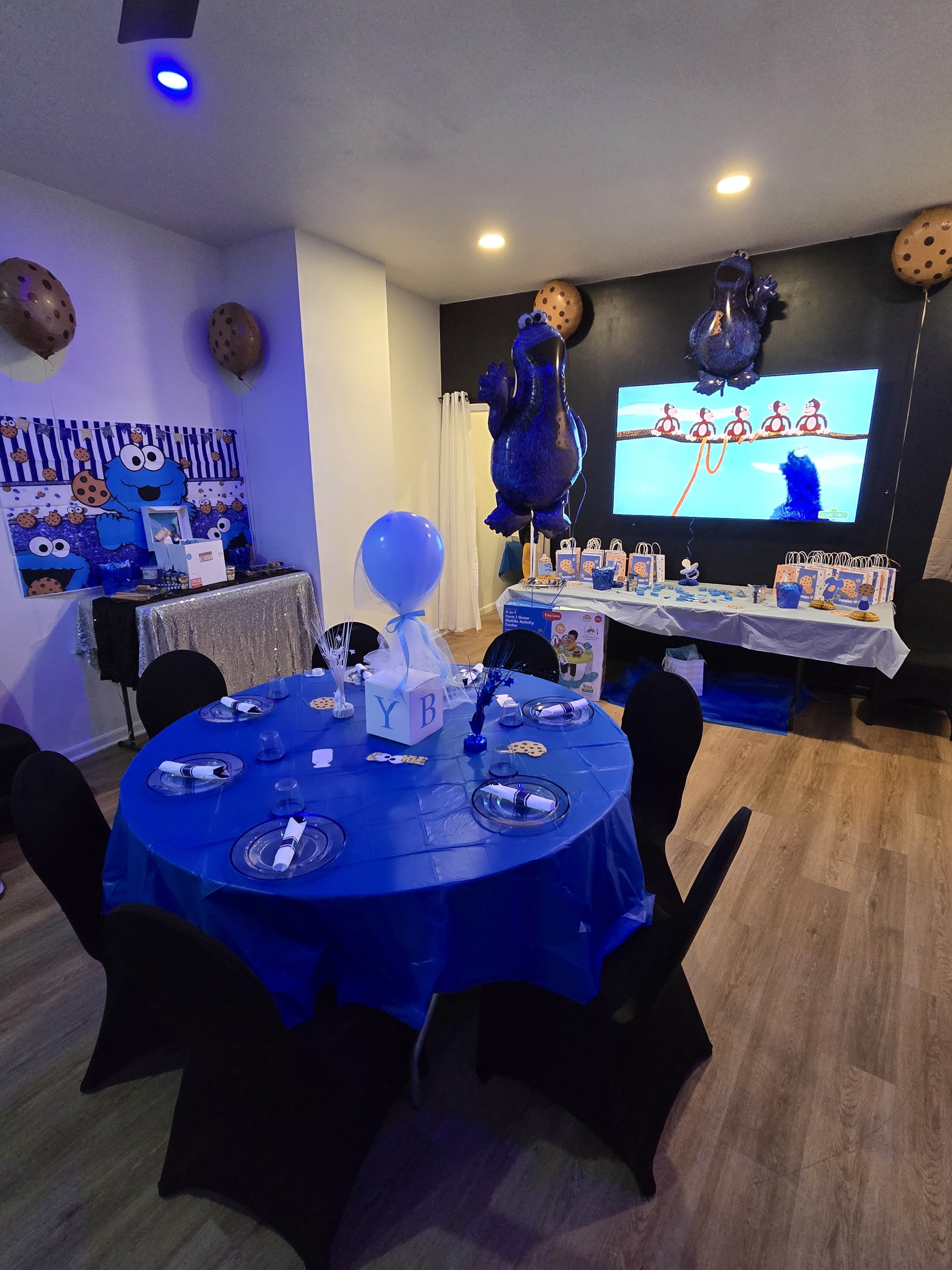 The scene depicts a decorated party room featuring a blue table with black chairs, cookie-themed decorations, and a large screen displaying an animated character.