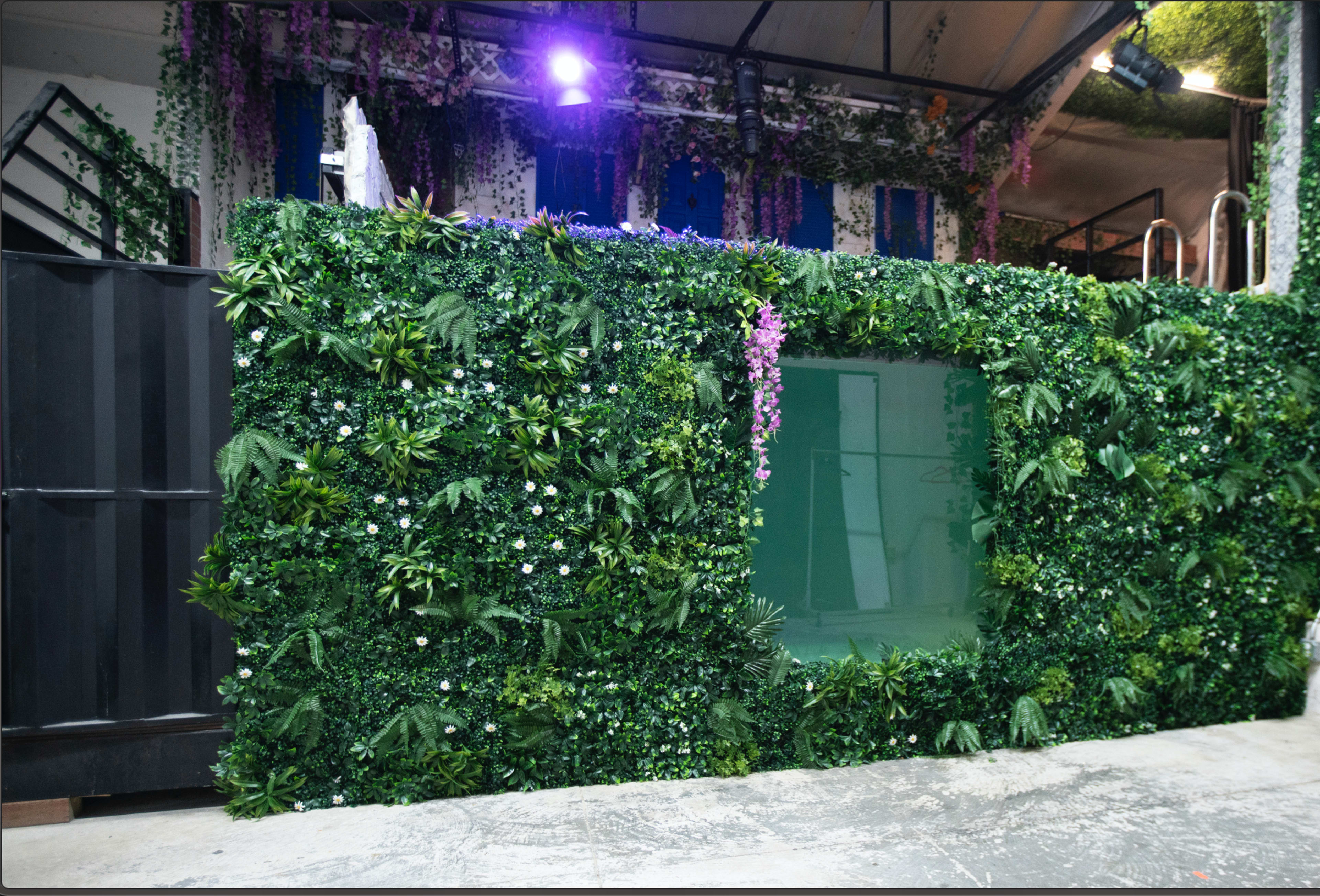 A wall covered in artificial greenery with flowers and a transparent window is shown in an indoor setting.