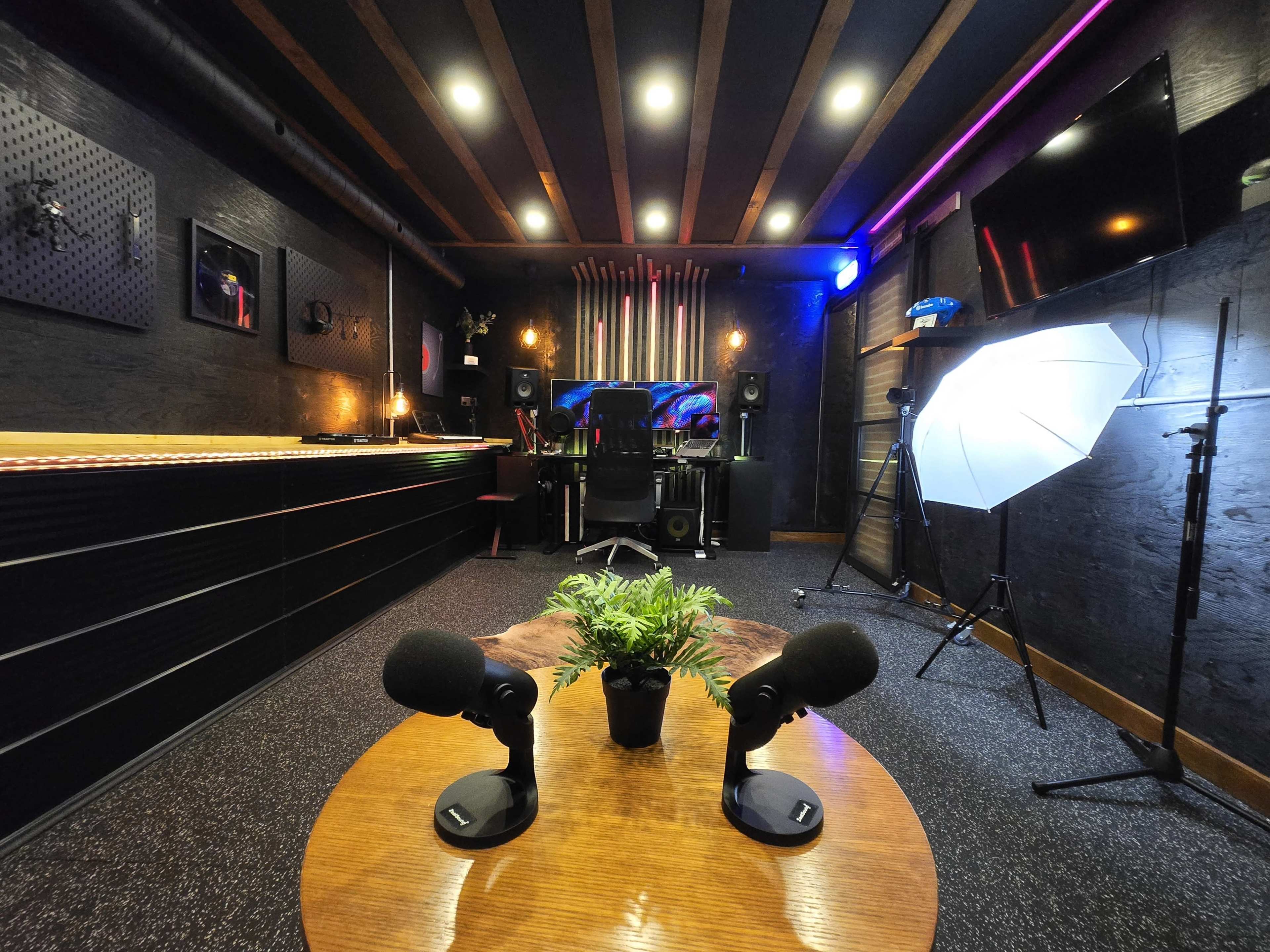 The image shows a modern recording studio featuring a wooden table with two microphones, a black chair, sound equipment, and ambient lighting.