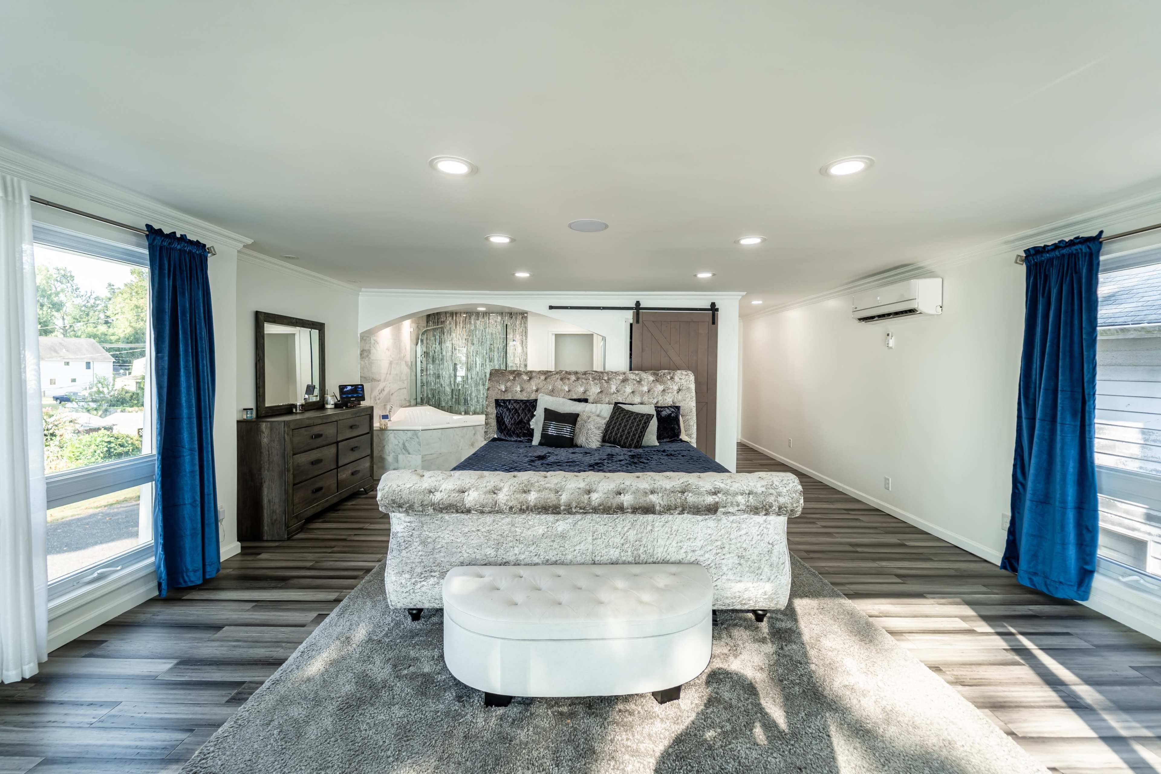 A spacious, modern bedroom featuring a large bed with a tufted headboard, a dresser, a mirror, and large windows with blue curtains.
