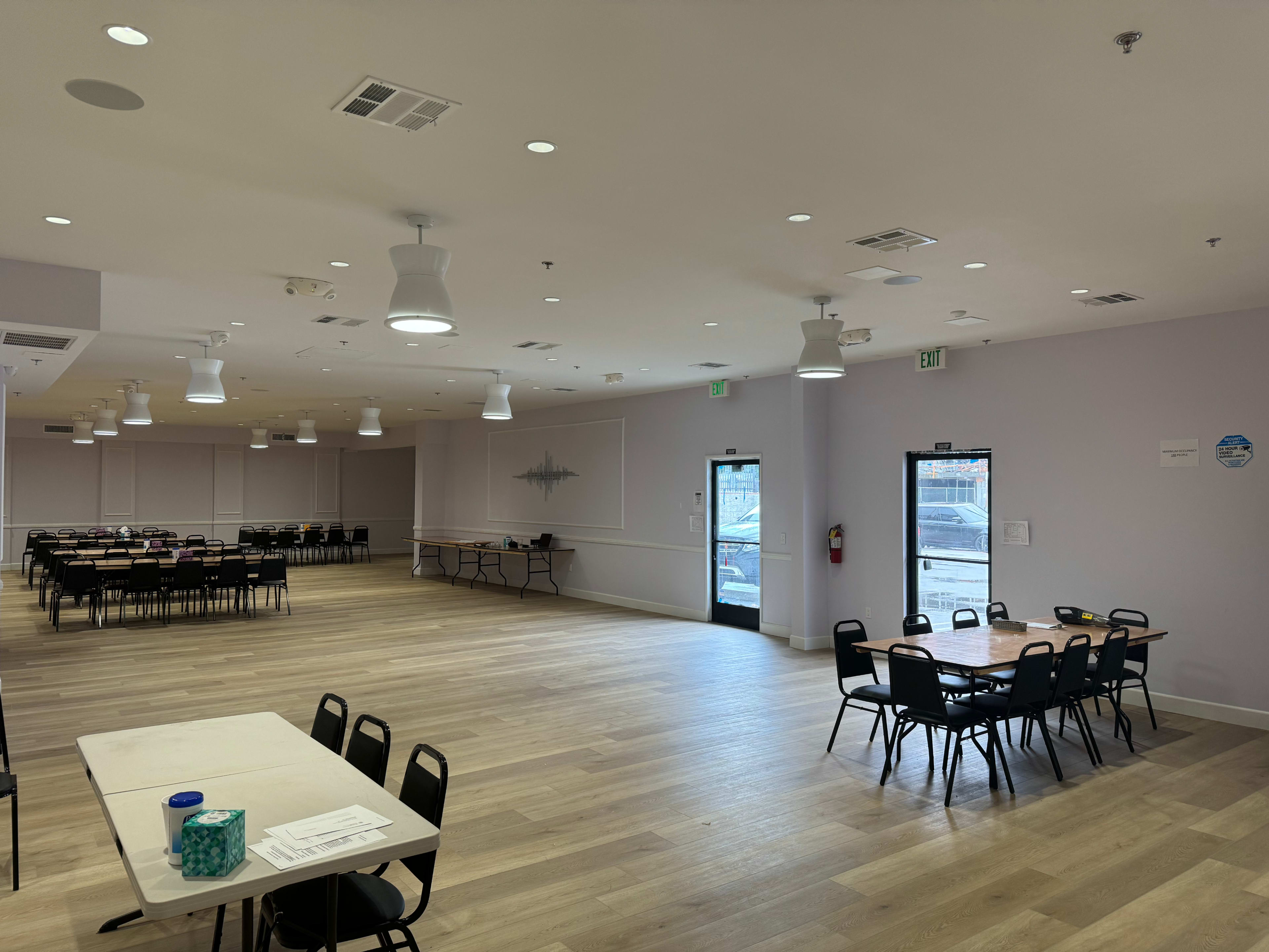 A spacious, well-lit room features multiple dining tables and chairs arranged for an event, with doors leading outside.