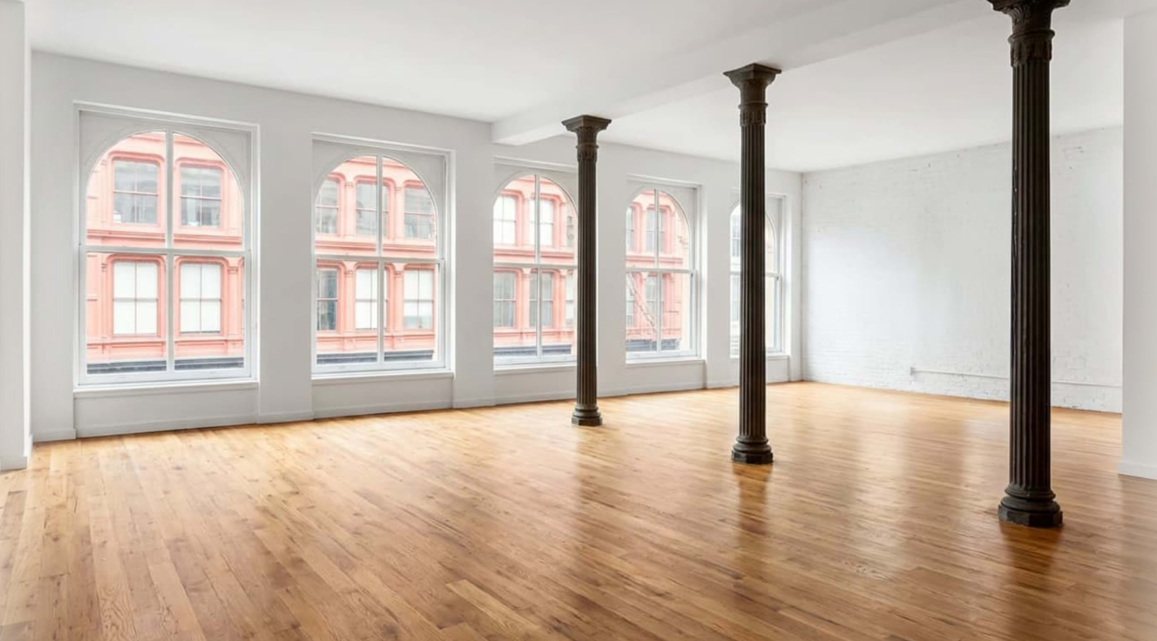 The image shows an empty, well-lit room with wooden floors, large windows featuring arched tops, and exposed columns.