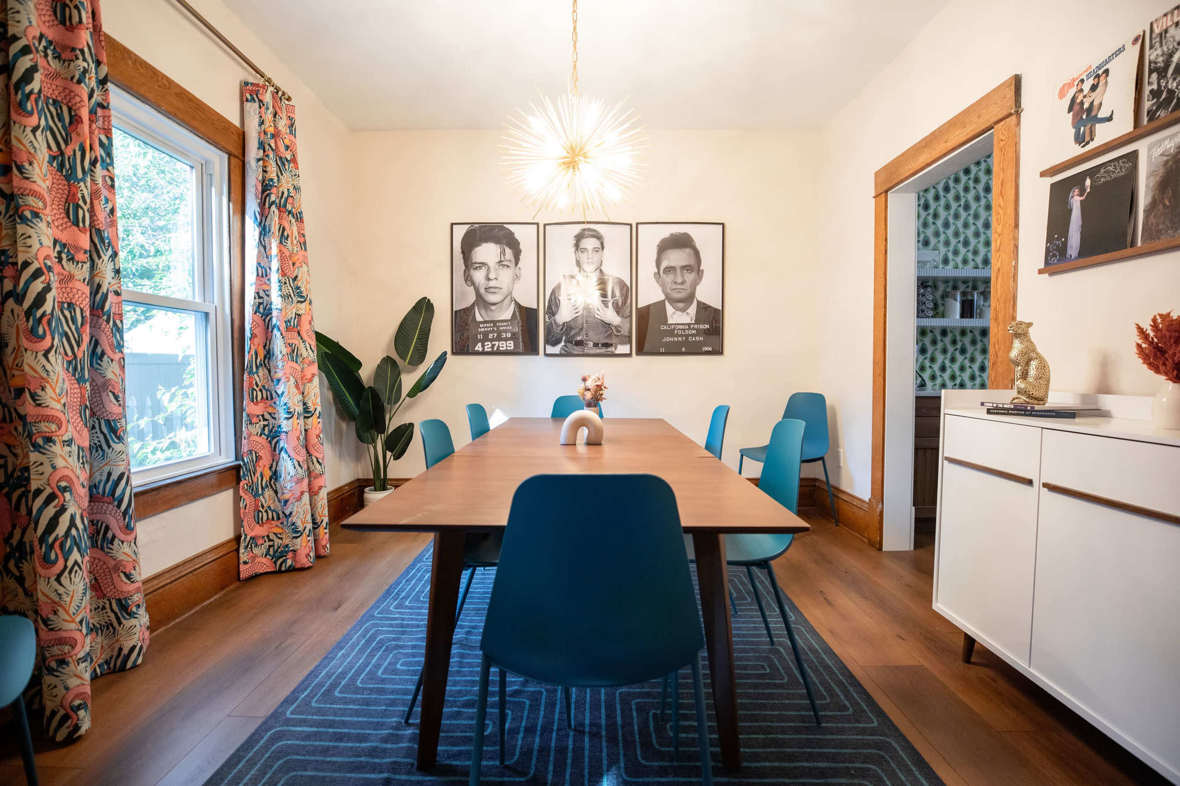 A dining room features a wooden table surrounded by blue chairs, with two framed portraits on the wall and a large light fixture overhead.