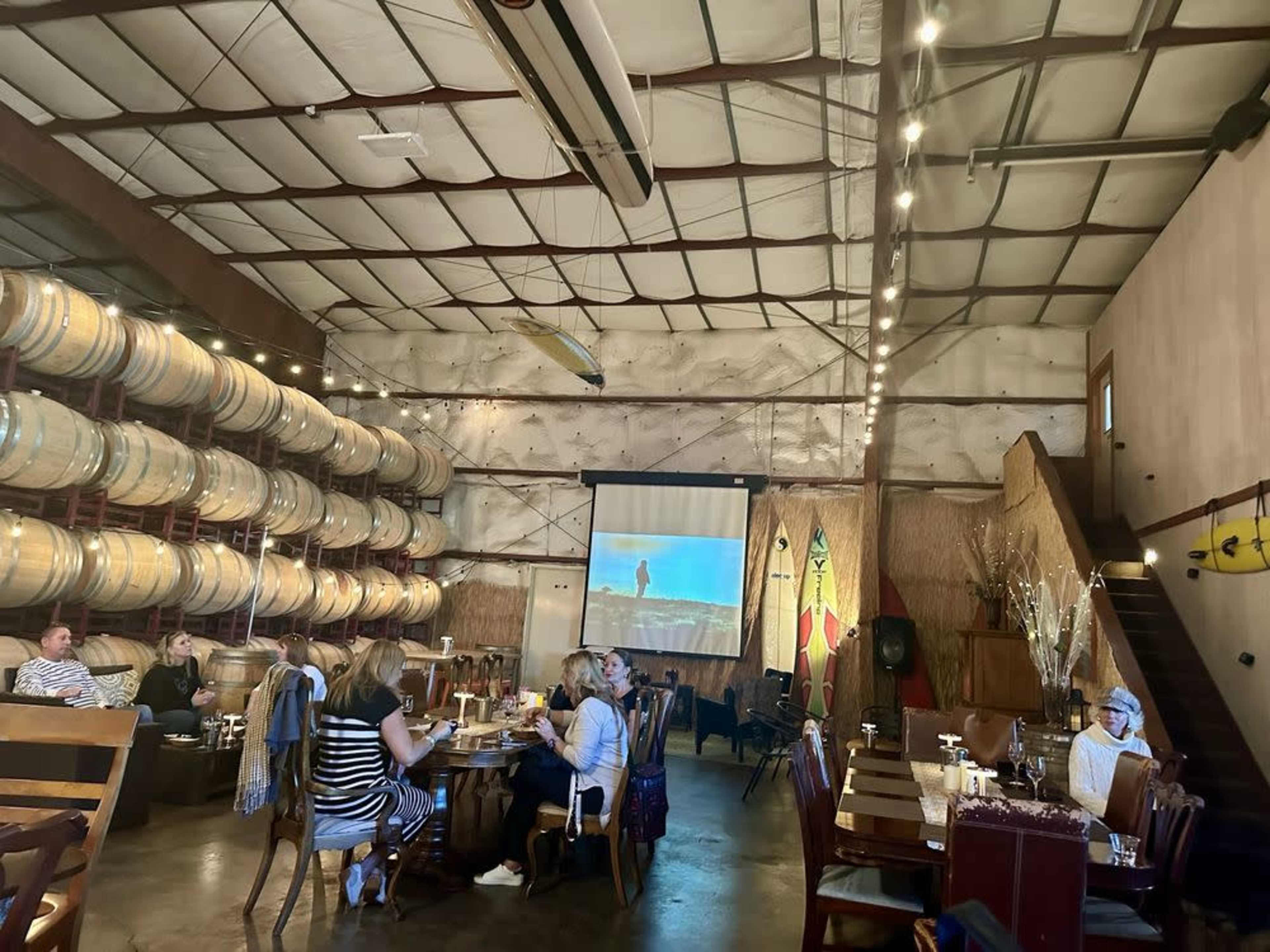 A group of people sits at tables in a rustic dining space, with wine barrels lining one wall and a projector screen displaying an image at the front.