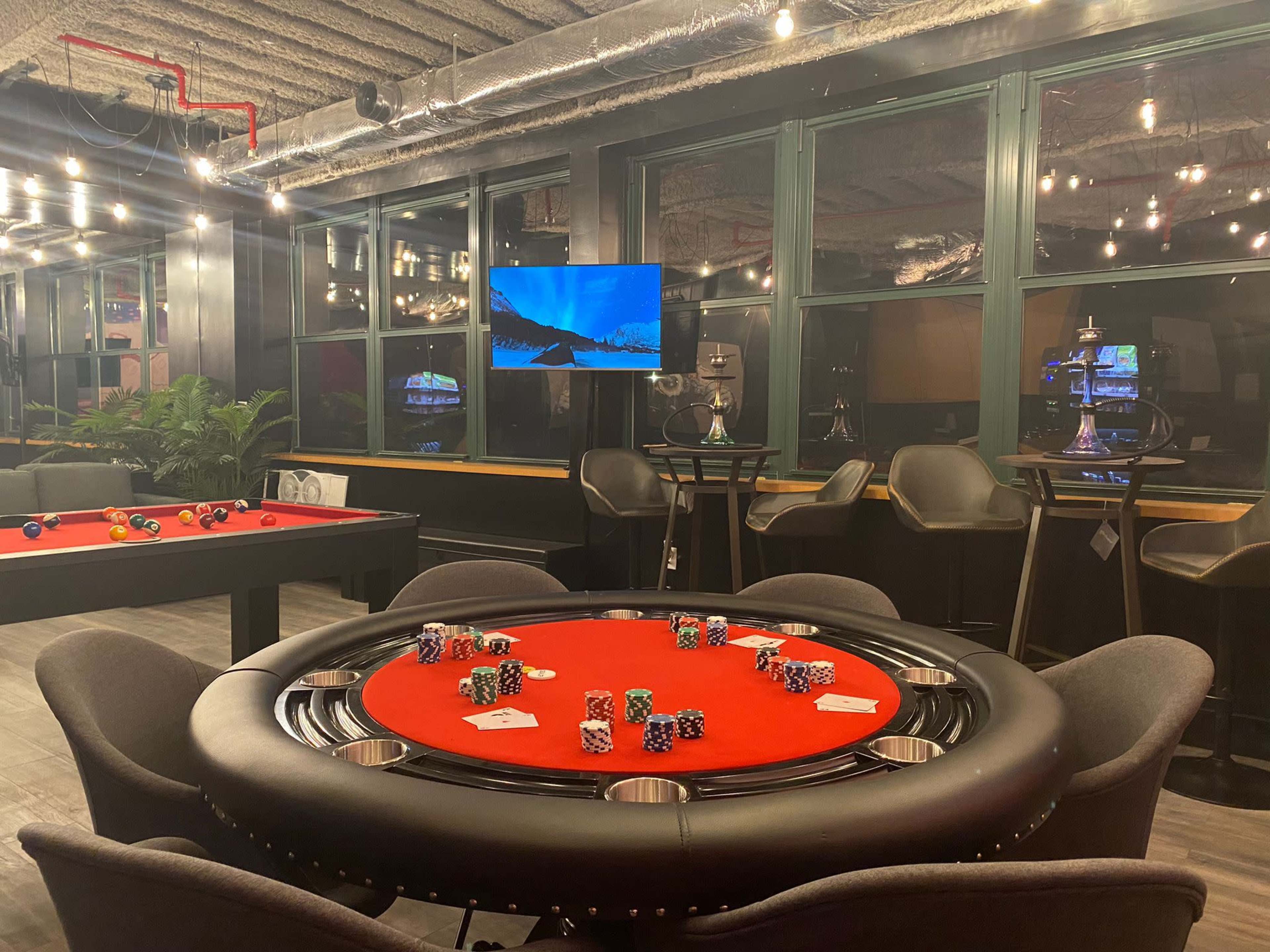 A poker table with chips arranged on a red felt surface, surrounded by modern seating and a television displaying a scenic image in a well-lit room.