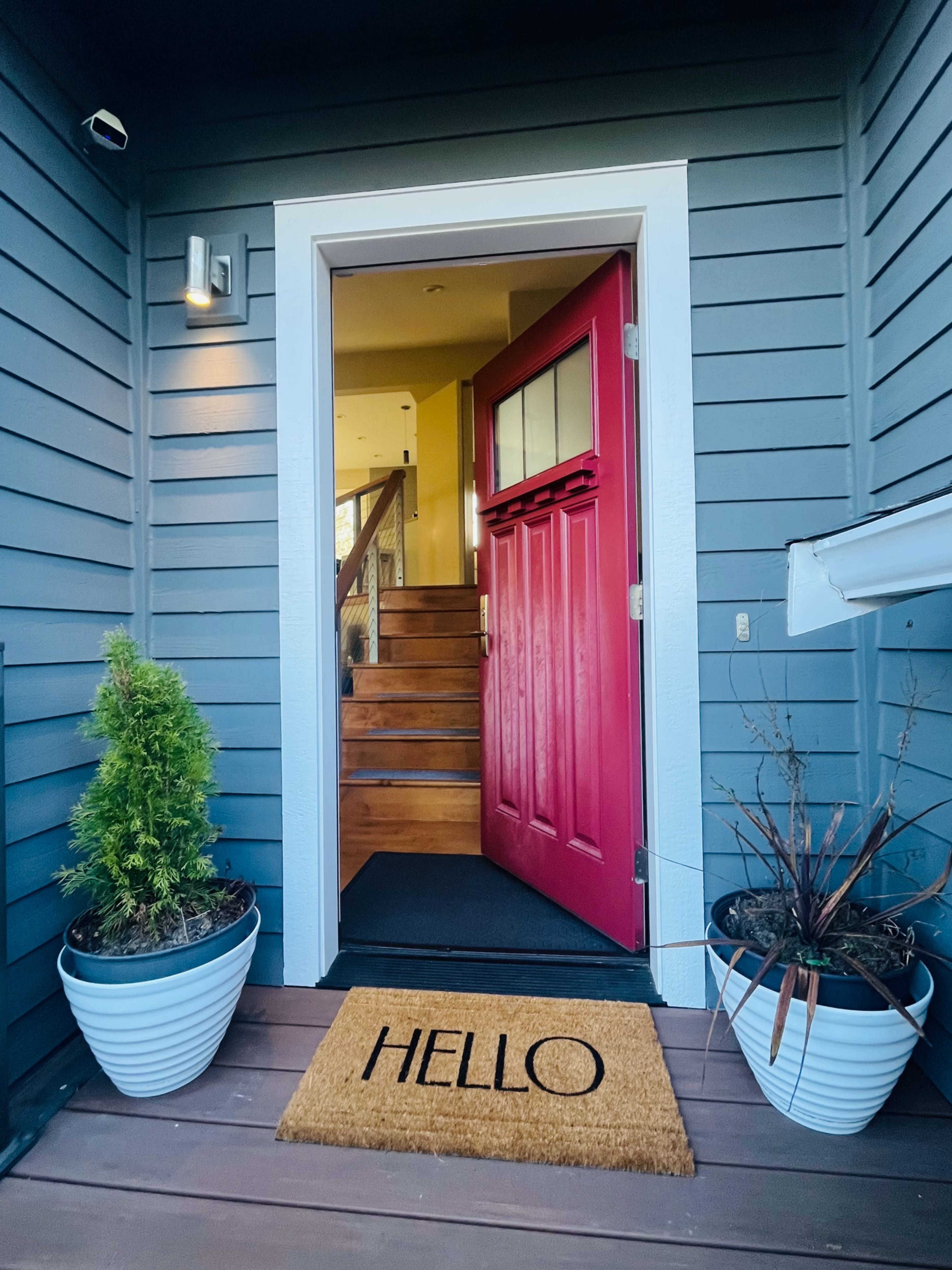 The image shows a brightly painted red front door open to a porch, flanked by two decorative planters and a welcome mat that reads "HELLO."