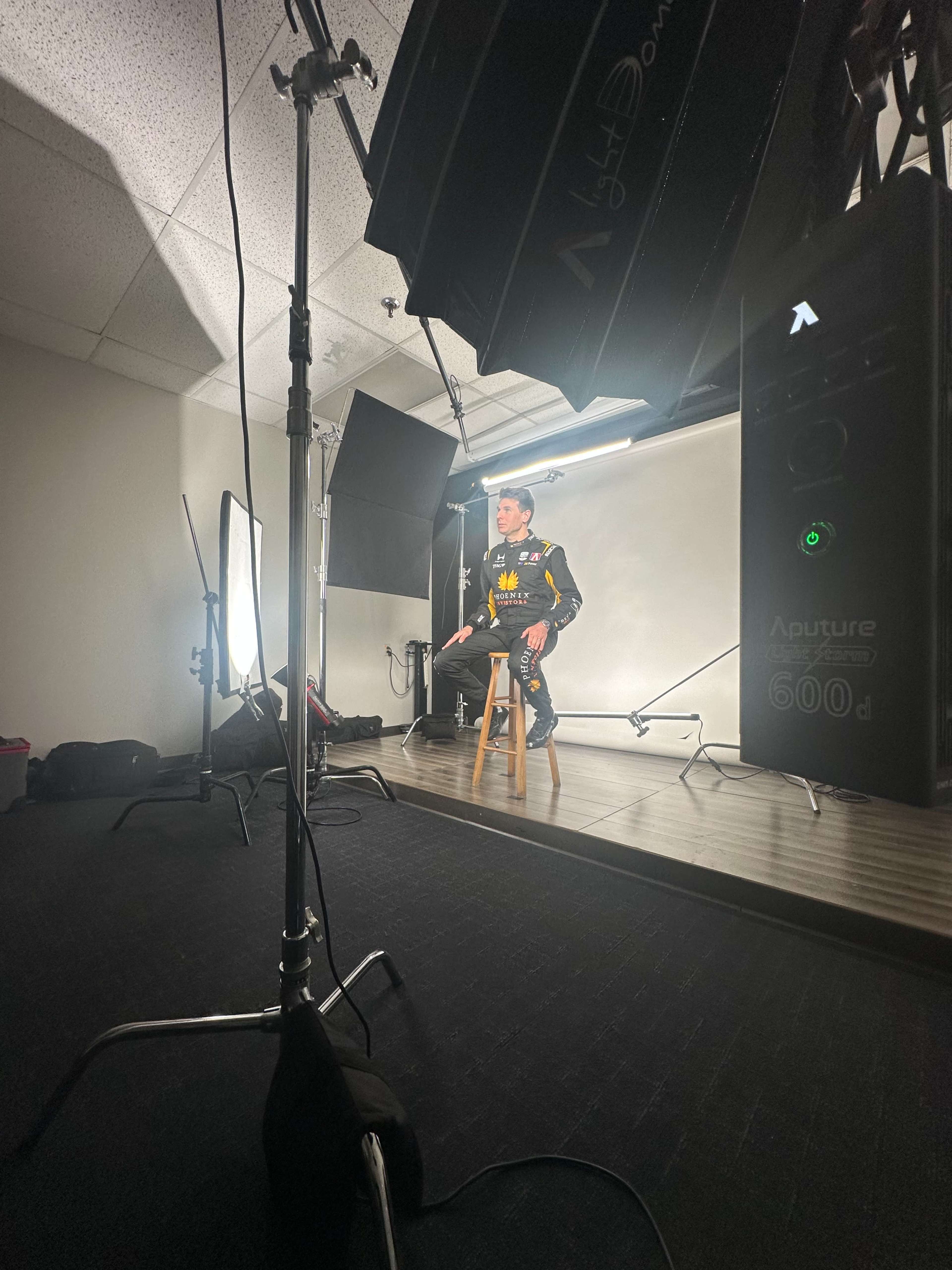 A person sits on a stool in a photography studio surrounded by various lighting equipment.
