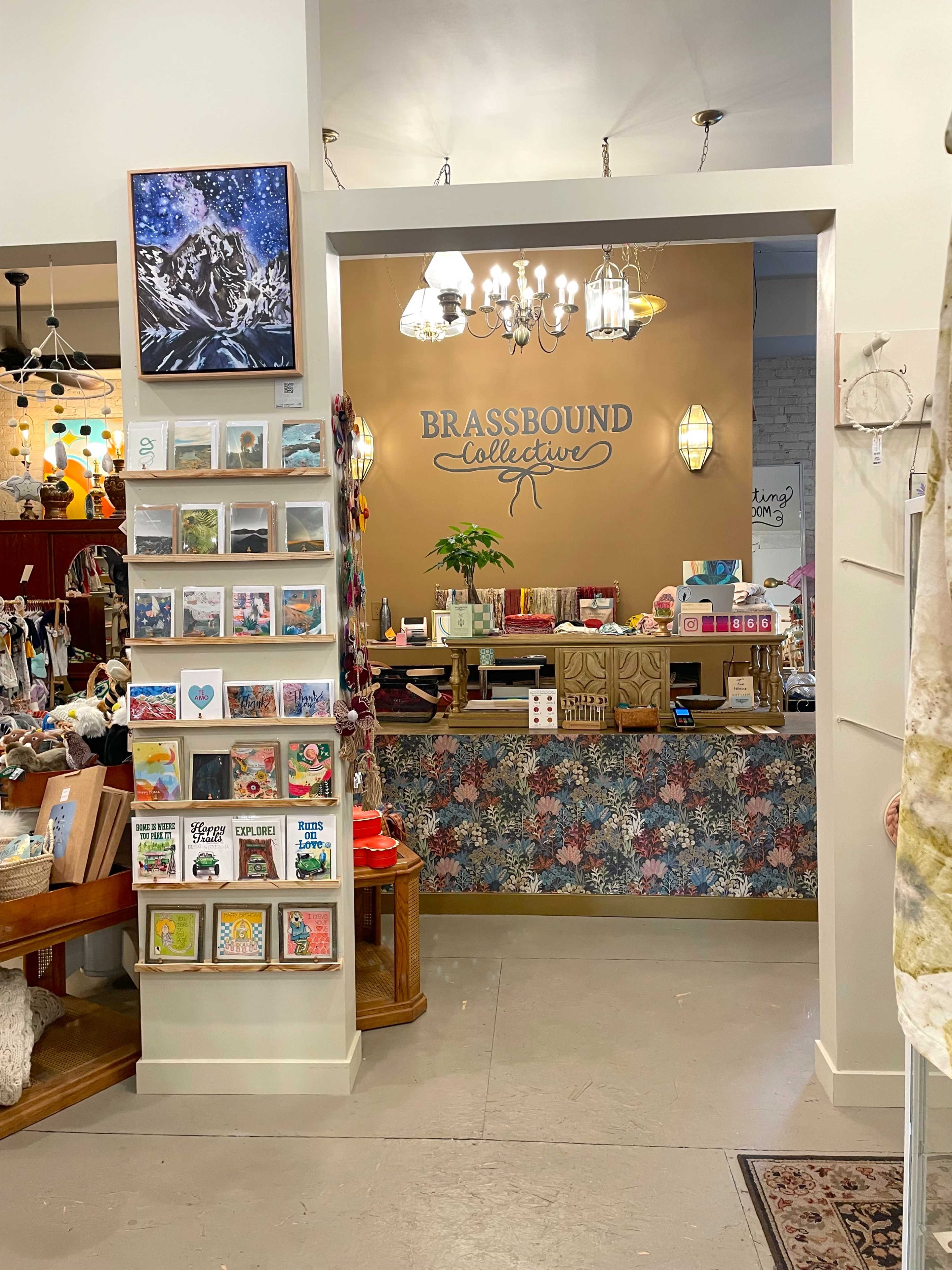The image shows the interior of a shop called "Brassbound Collective," featuring a display of greeting cards on the left and a wooden counter with various products in the background.