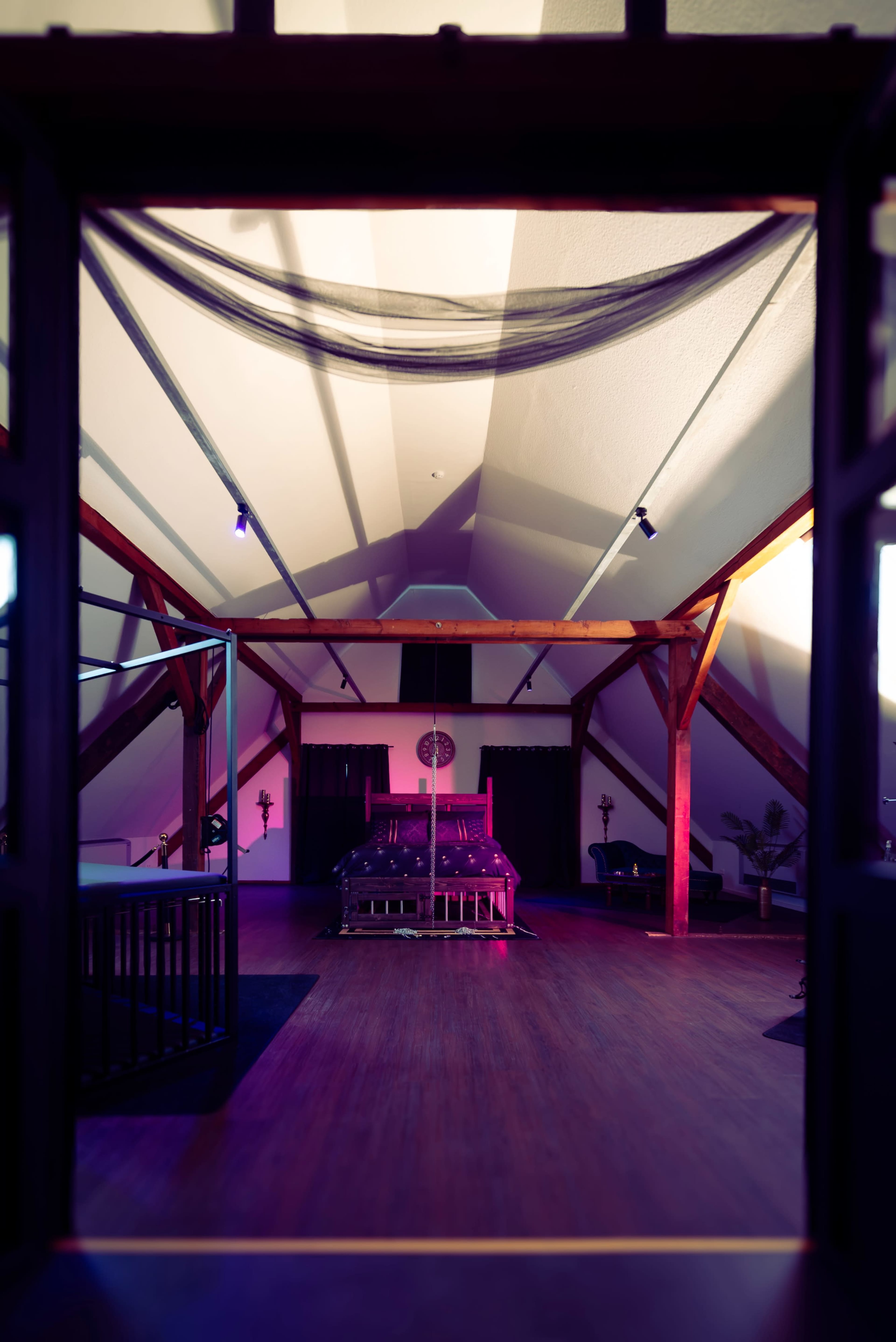 The image shows a spacious room with high wooden beams, featuring a centrally placed bed and various seating areas, illuminated by colorful lights.