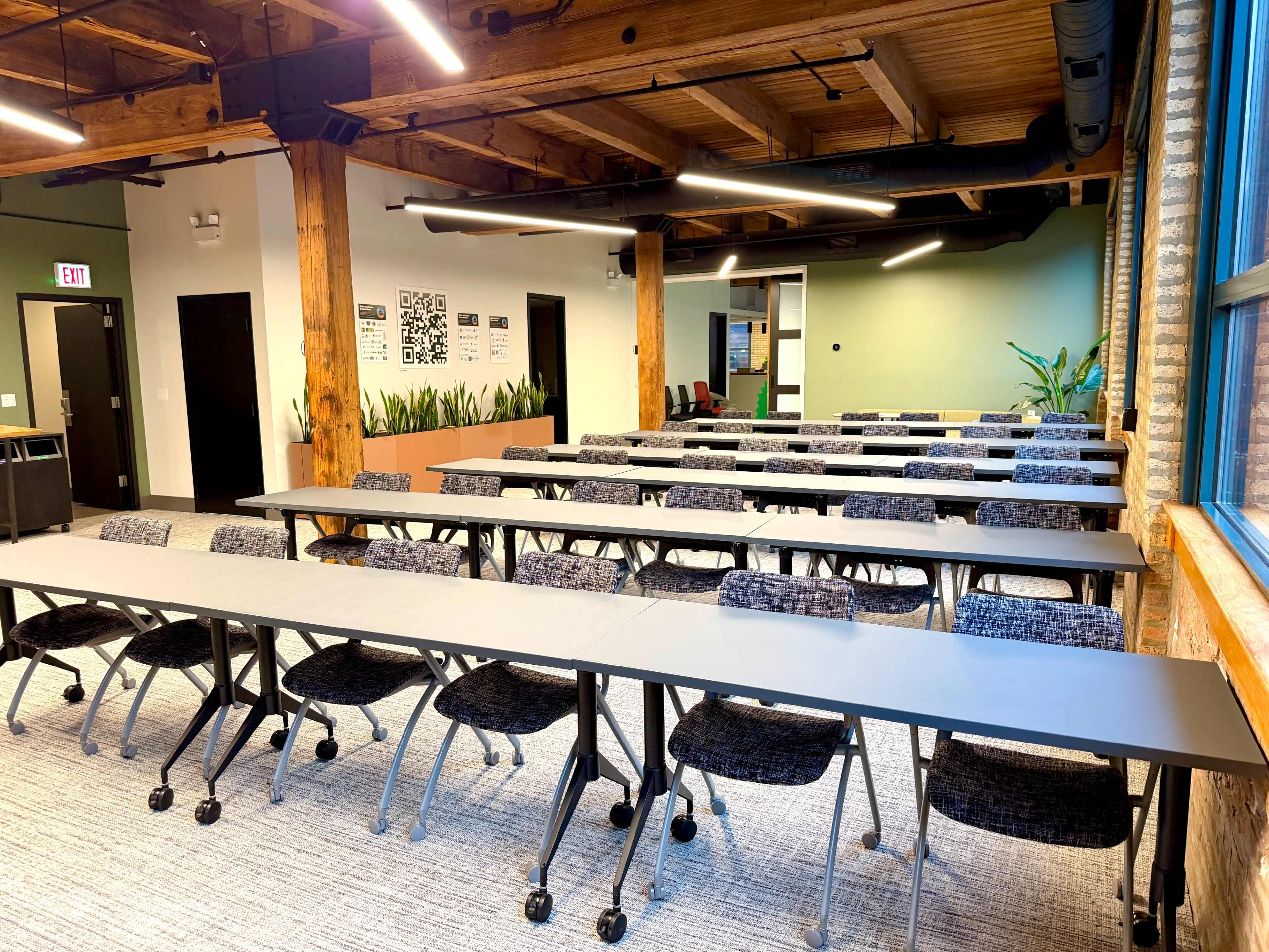 The image shows a modern classroom or meeting space with rows of tables and chairs arranged neatly, featuring wooden beams and plants in the background.