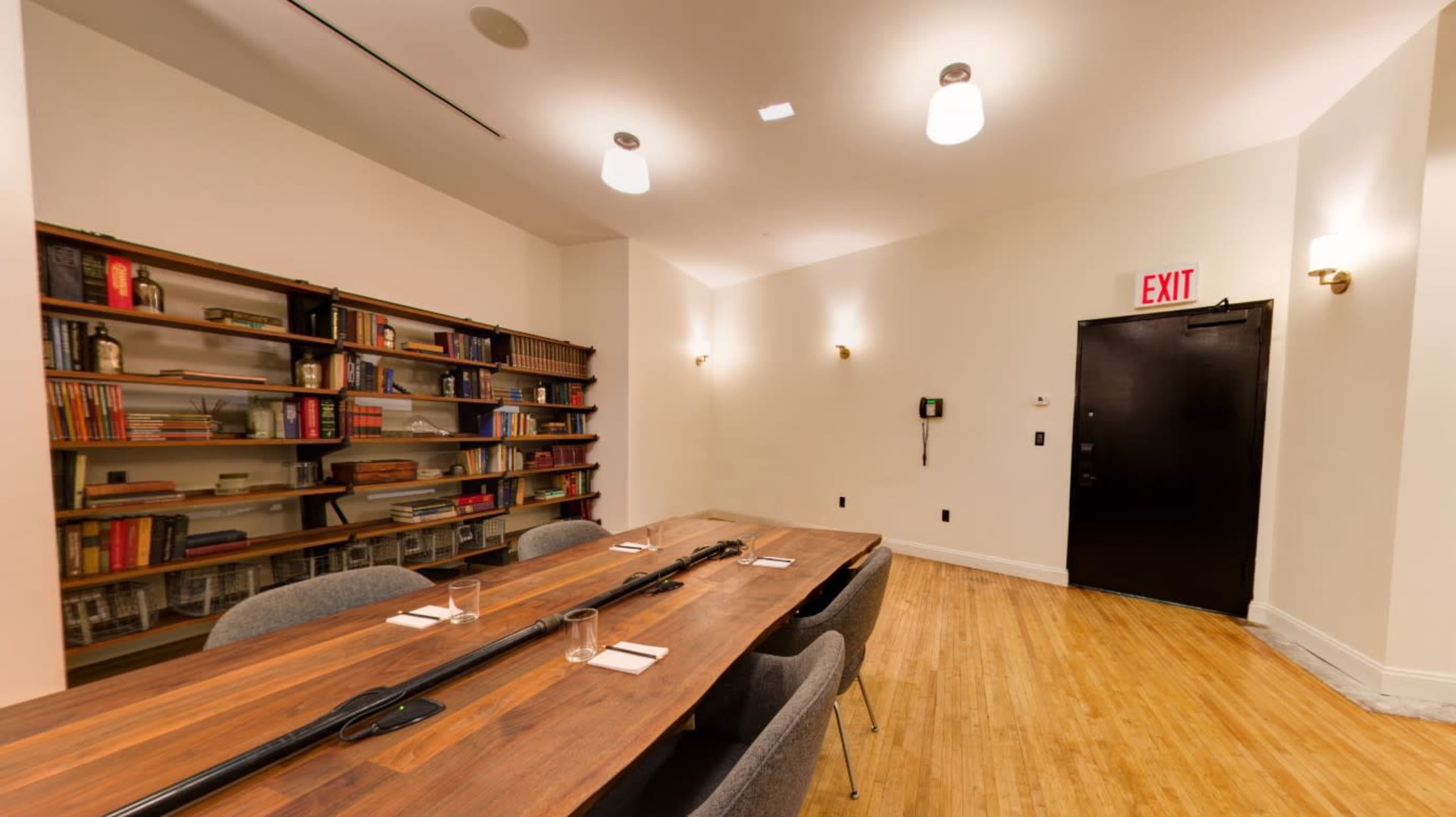 The room features a wooden table surrounded by chairs, a bookshelf filled with books, and a black exit door.