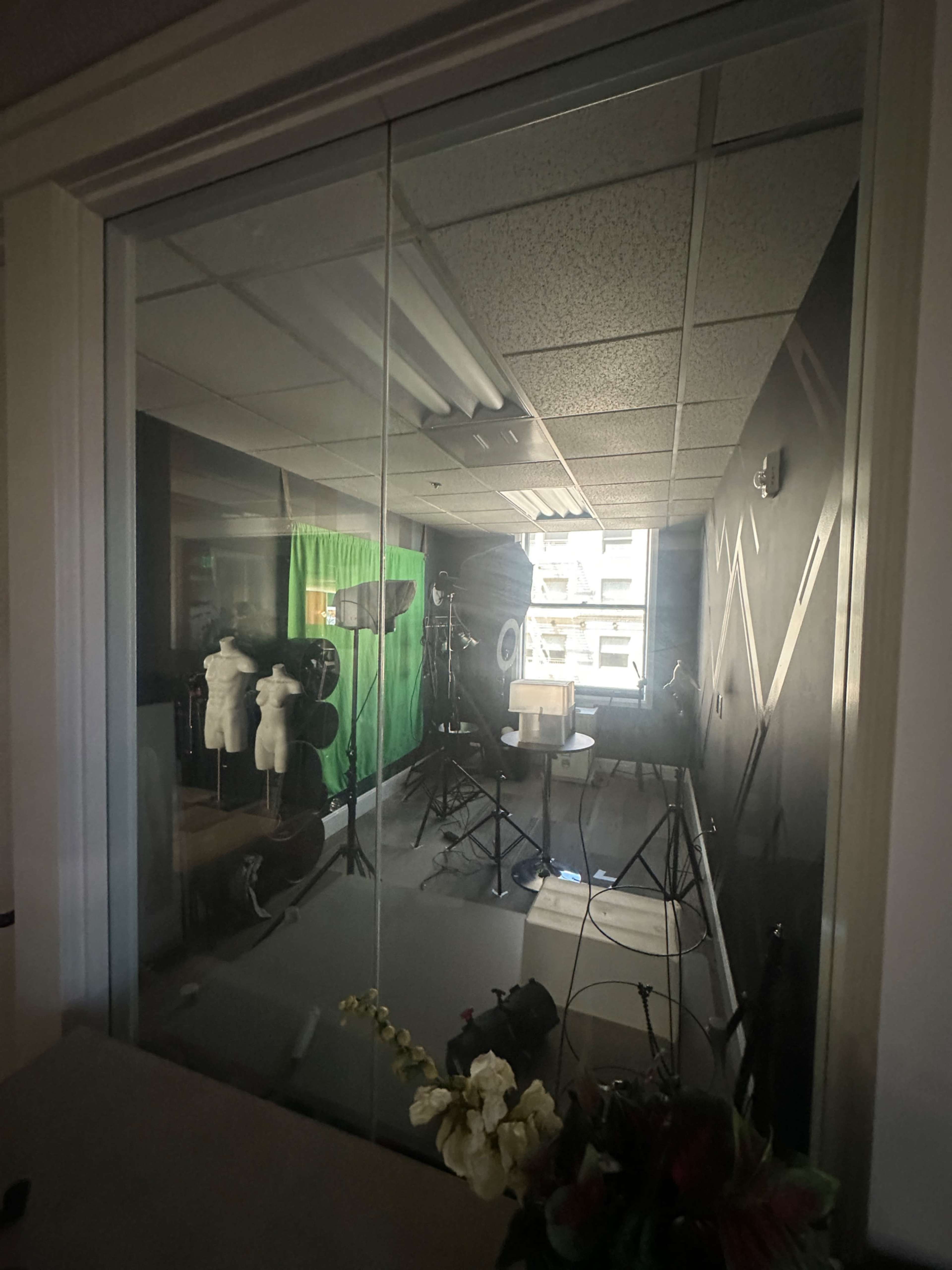 The image shows a studio space visible through a glass wall, featuring a green backdrop, lighting equipment, and various props arranged inside.