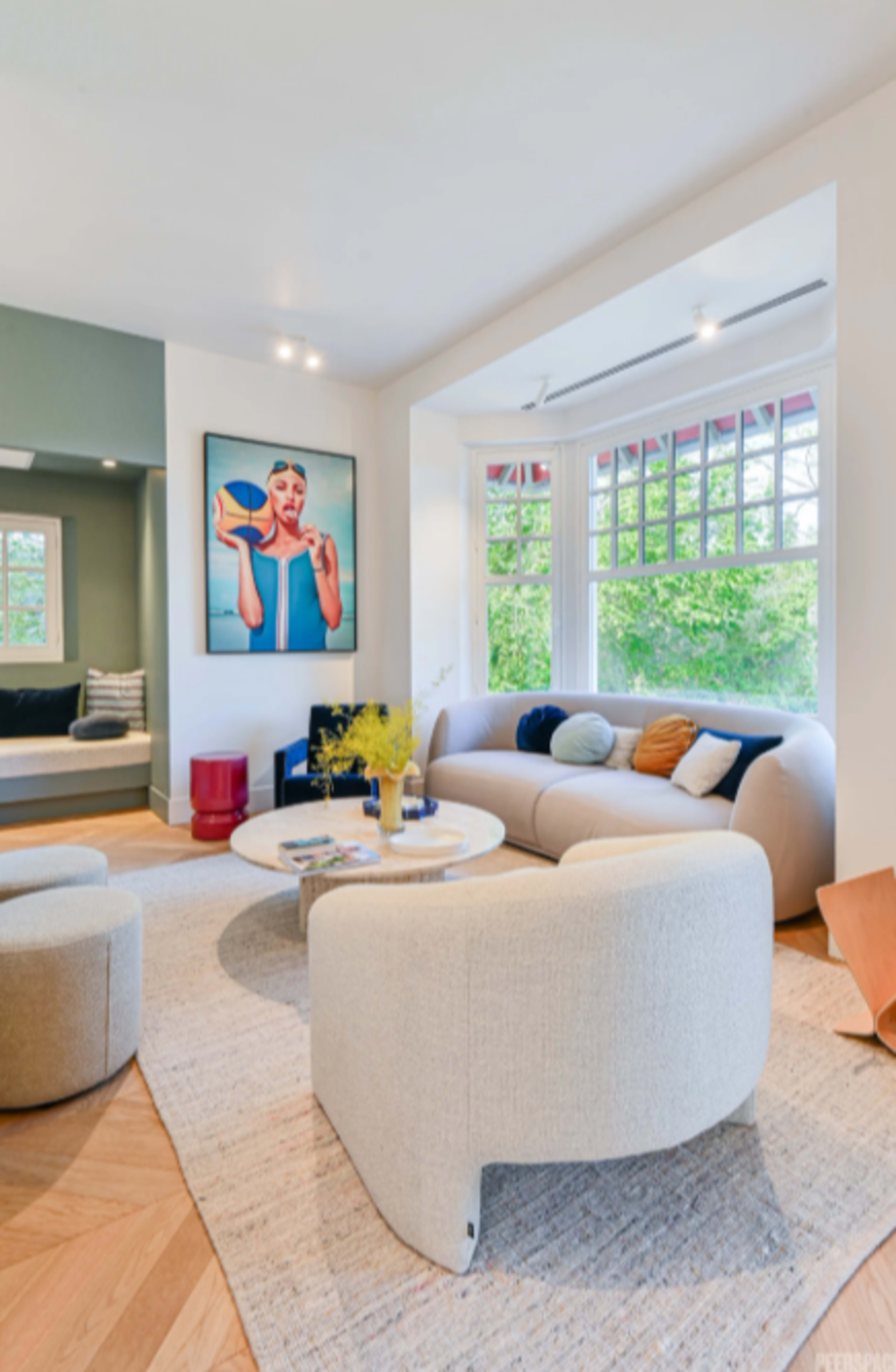 The image shows a modern living room featuring a curved sofa, a round coffee table, and a large art piece on the wall, with natural light coming in from large windows.