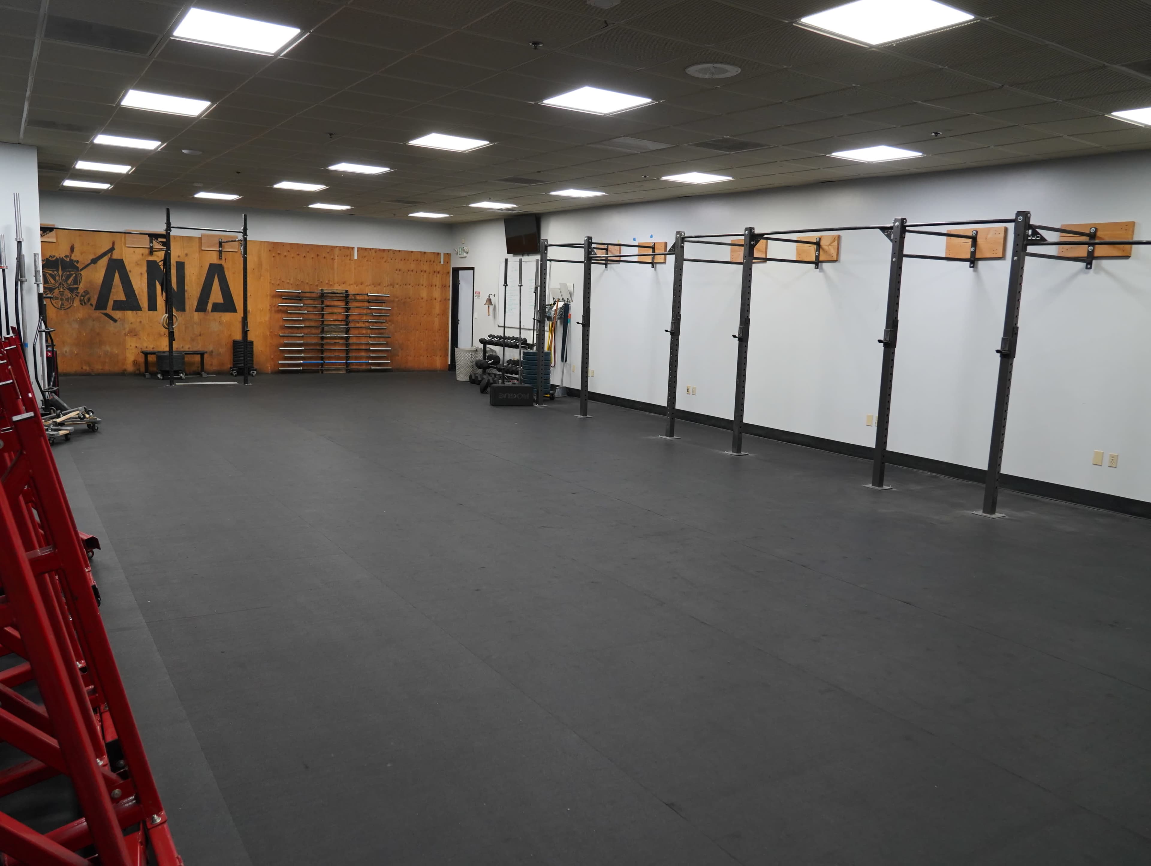 The image shows an empty gym space with black rubber flooring, wall-mounted bars, and a wooden wall with various exercise equipment organized on shelves.