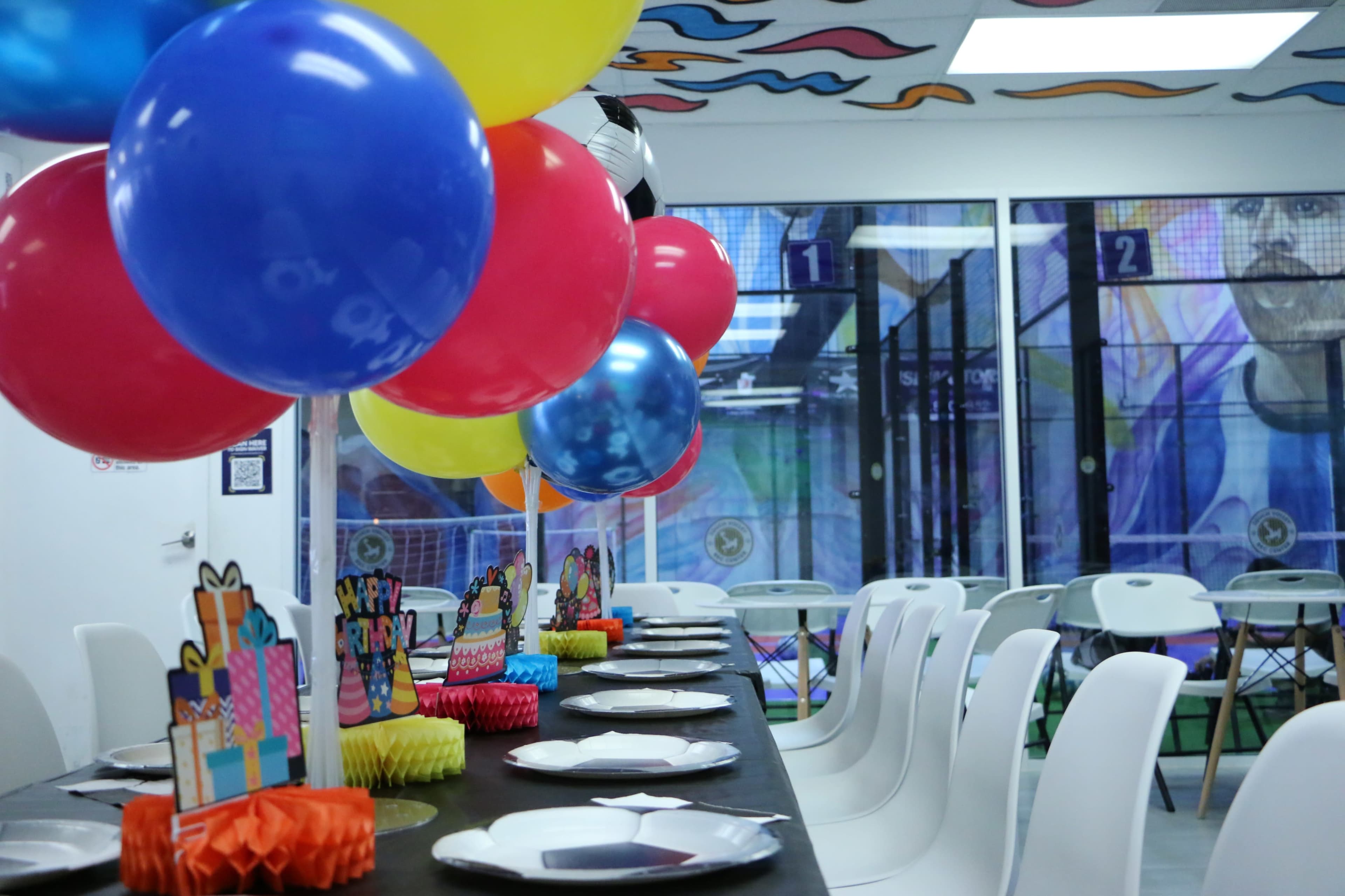 A festive party setup features a long table adorned with colorful balloons and decorative items, set against a backdrop of vibrant wall art and an indoor activity space.