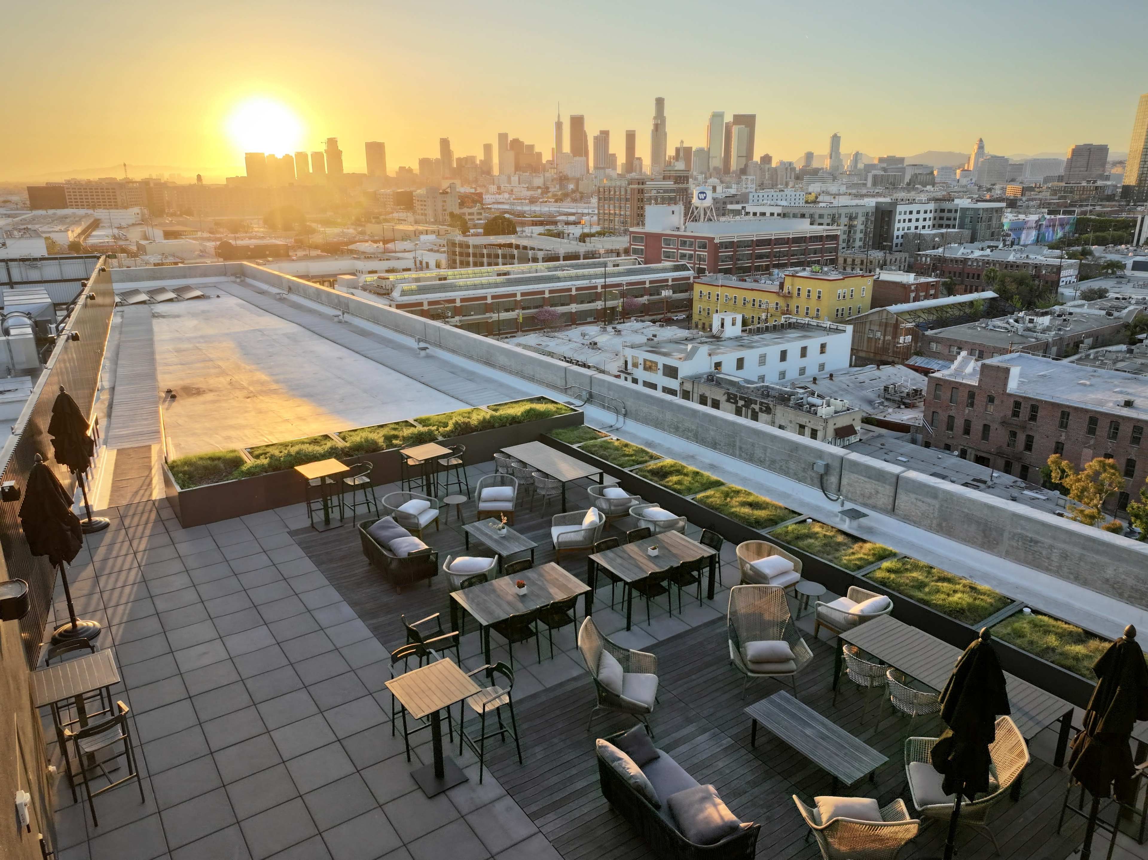 A rooftop terrace with wooden tables and chairs overlooking a city skyline at sunset.