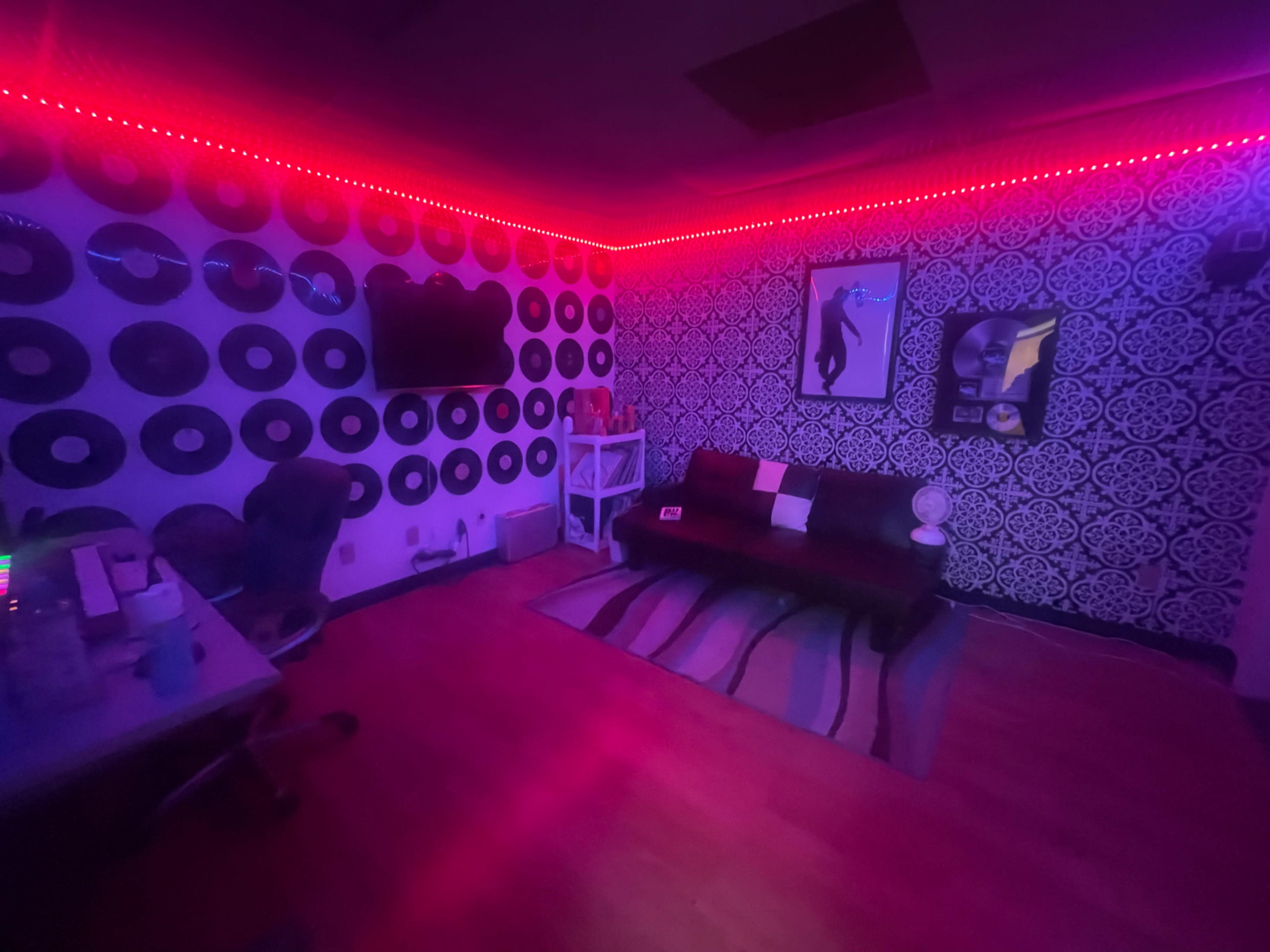 A room with purple and red lighting, adorned with wall decorations of vinyl records and a patterned wallpaper, featuring a couch and a small table with a television on the wall.