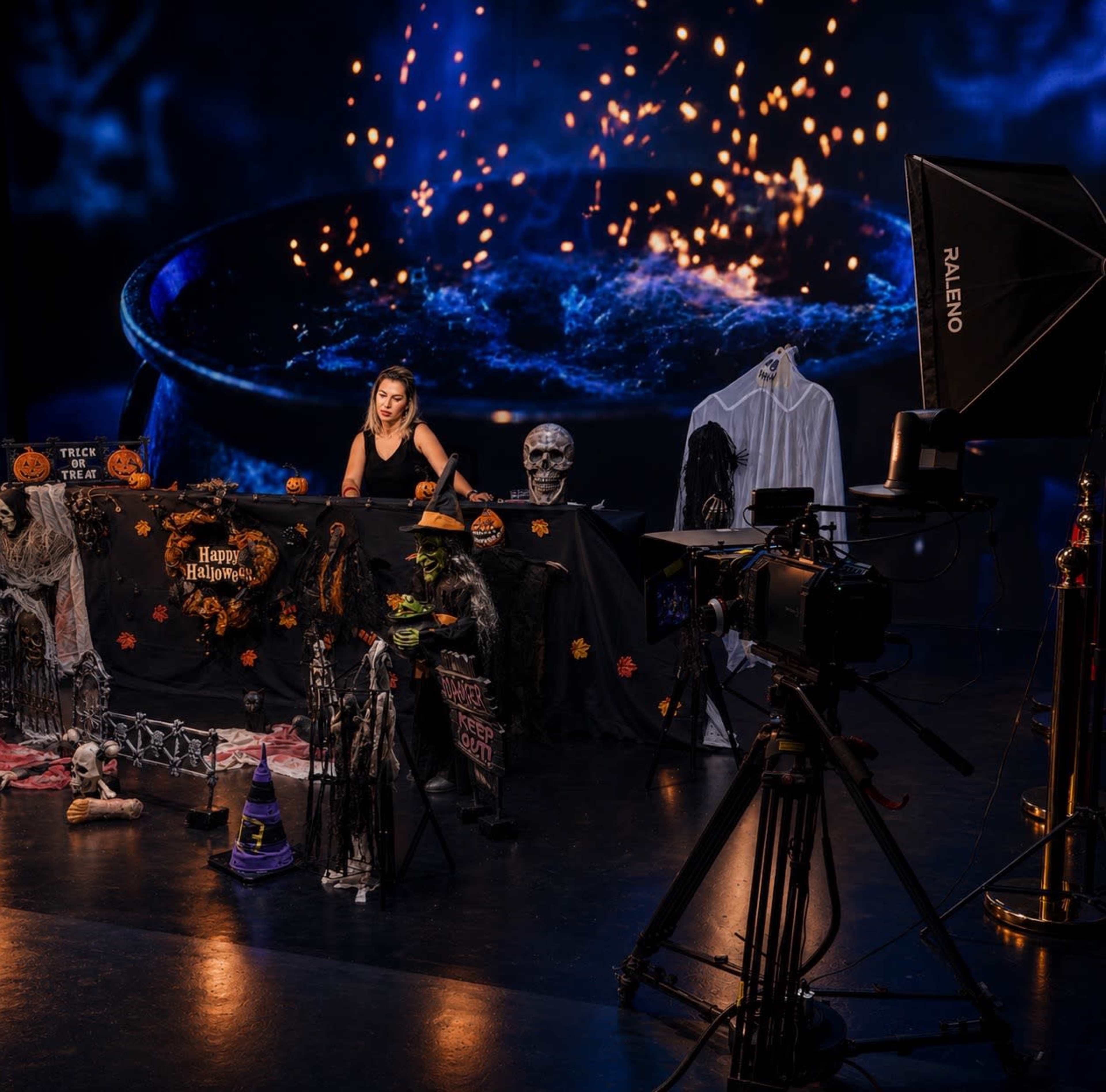 A woman sits behind a decorated table with Halloween-themed props and a camera setup in a studio filled with blue light and sparkles.
