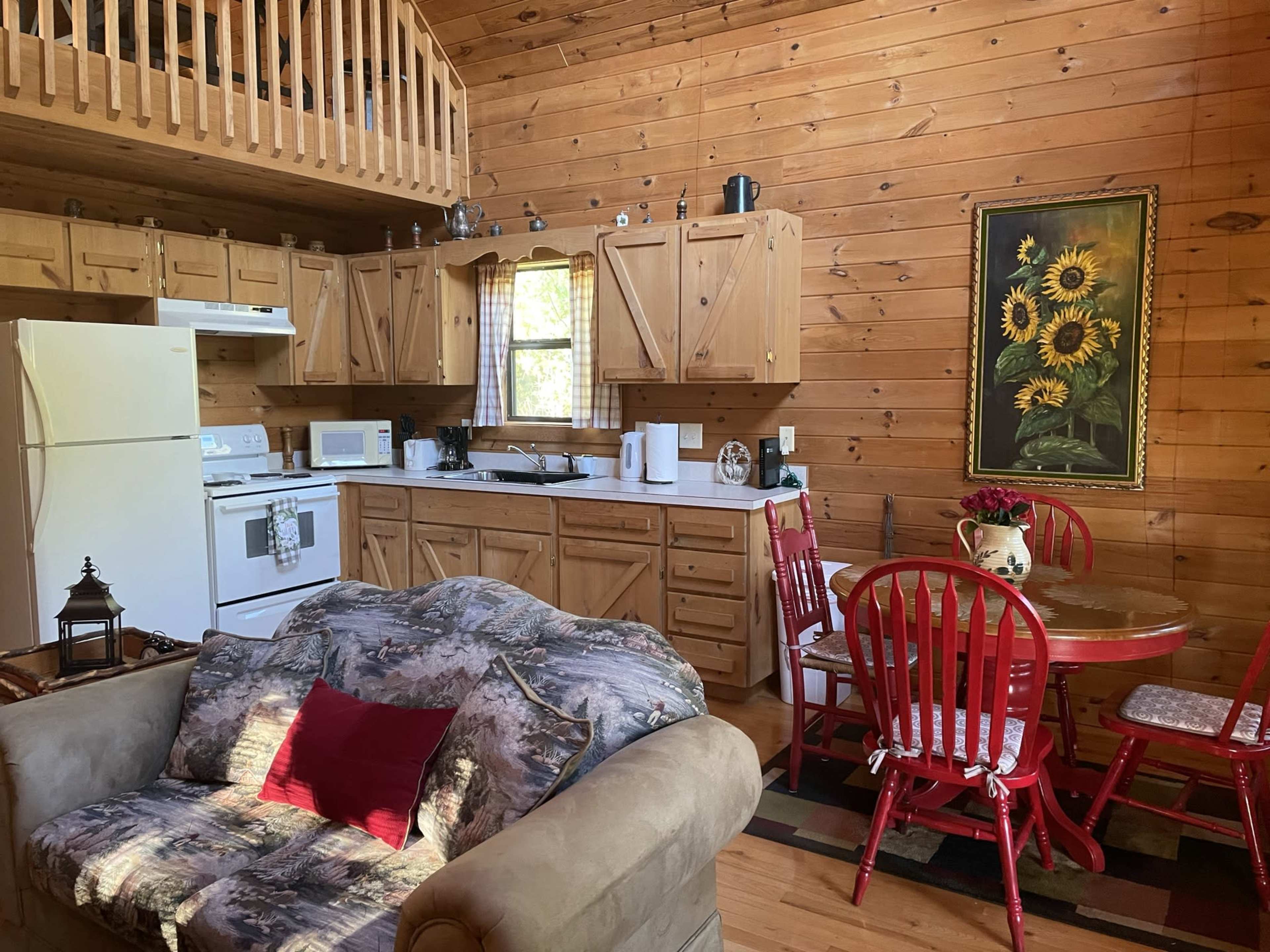 A wooden cabin interior features a kitchen area with pine cabinets, a white refrigerator, and a dining table with red chairs.