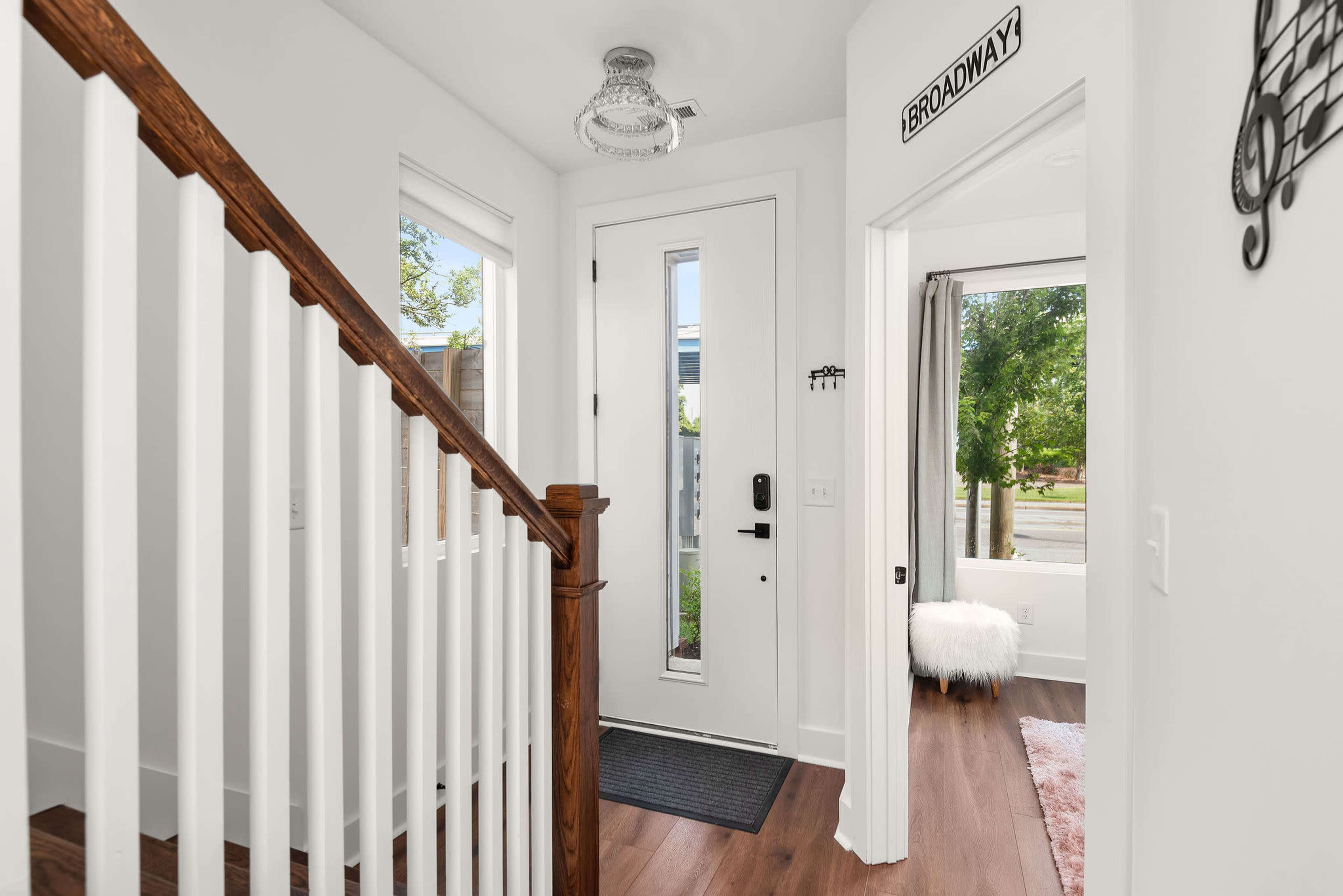 The image shows a well-lit entryway with a staircase on the left, a front door at the center, and a window with a view of the outdoors on the right.