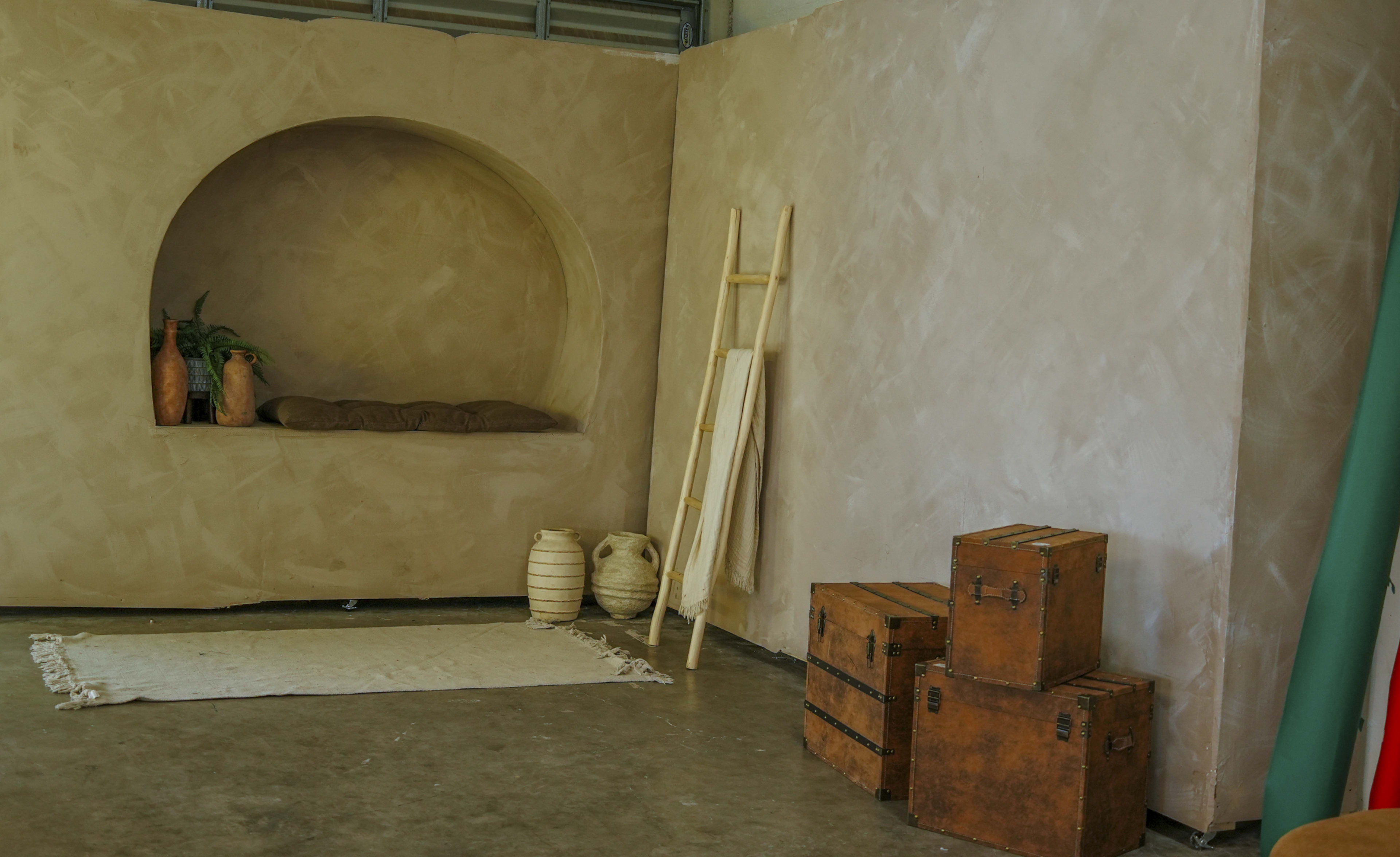 The image shows a minimalist interior space with a curved alcove featuring plants and cushions, a wooden ladder, woven textile on the floor, and stacked wooden boxes.