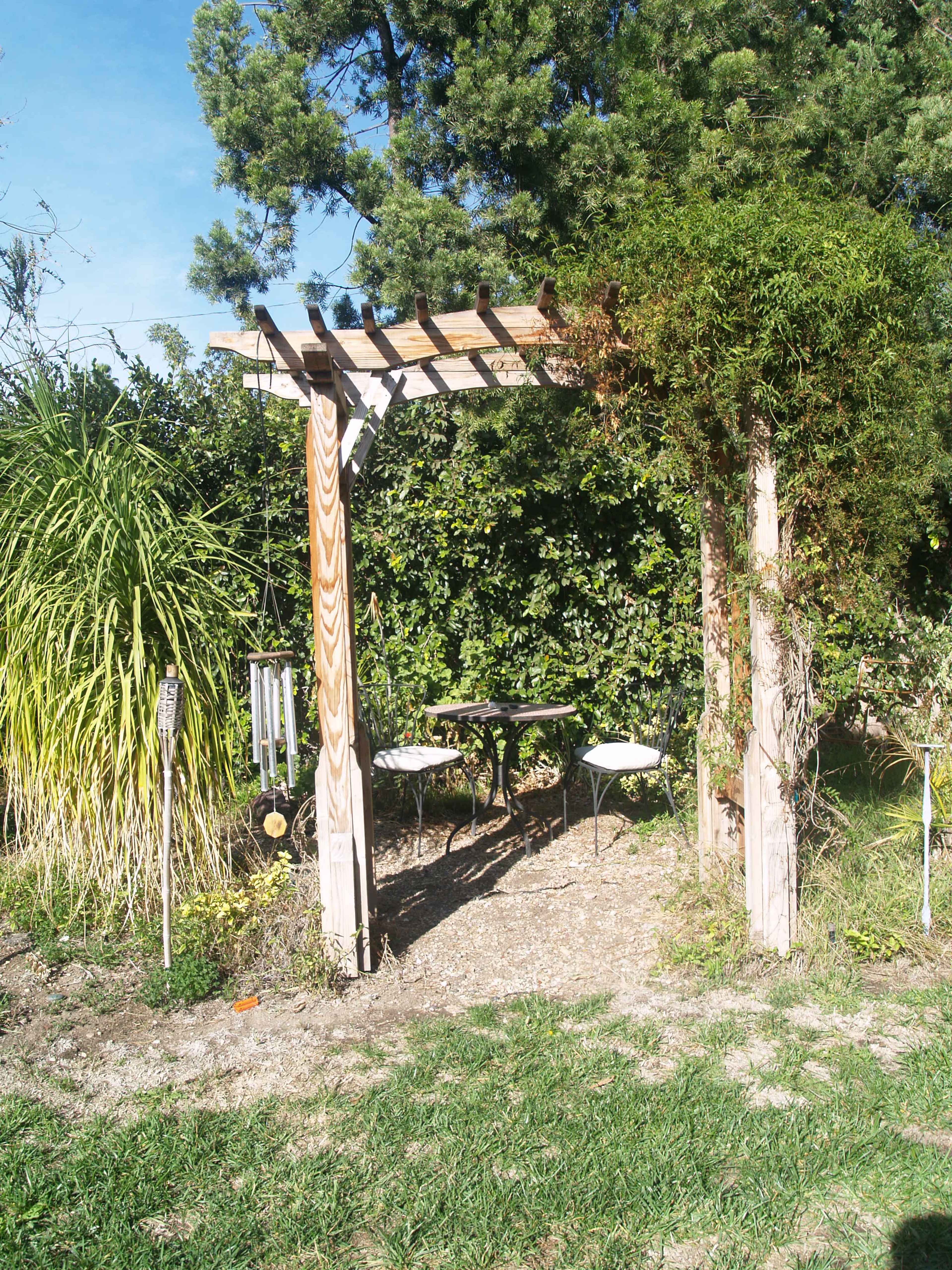 A wooden trellis leads to a small seating area with a table and chairs, surrounded by lush greenery.