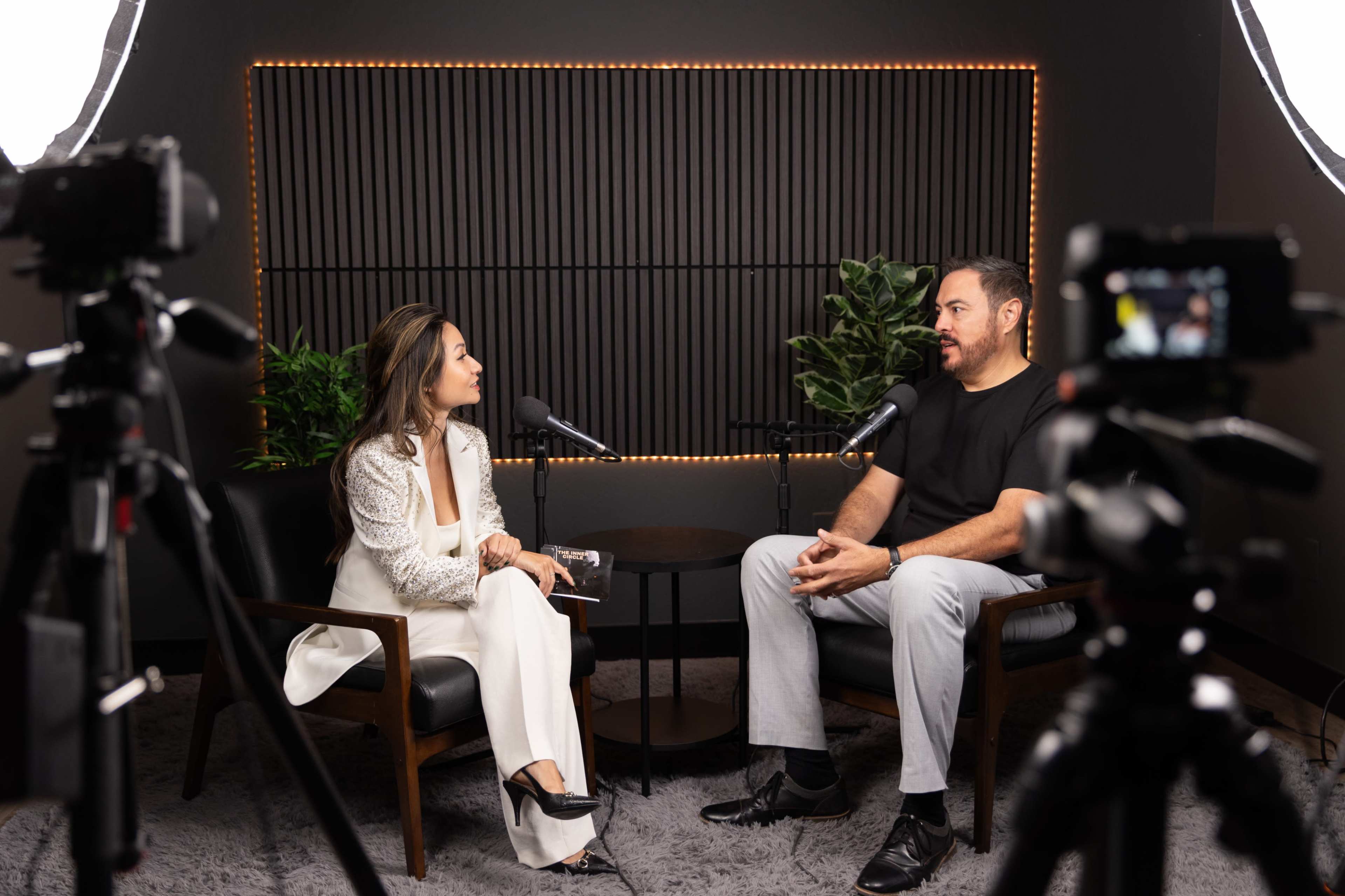 A woman and a man sit in a recorded interview setting, flanked by microphones and cameras, with plants in the background.