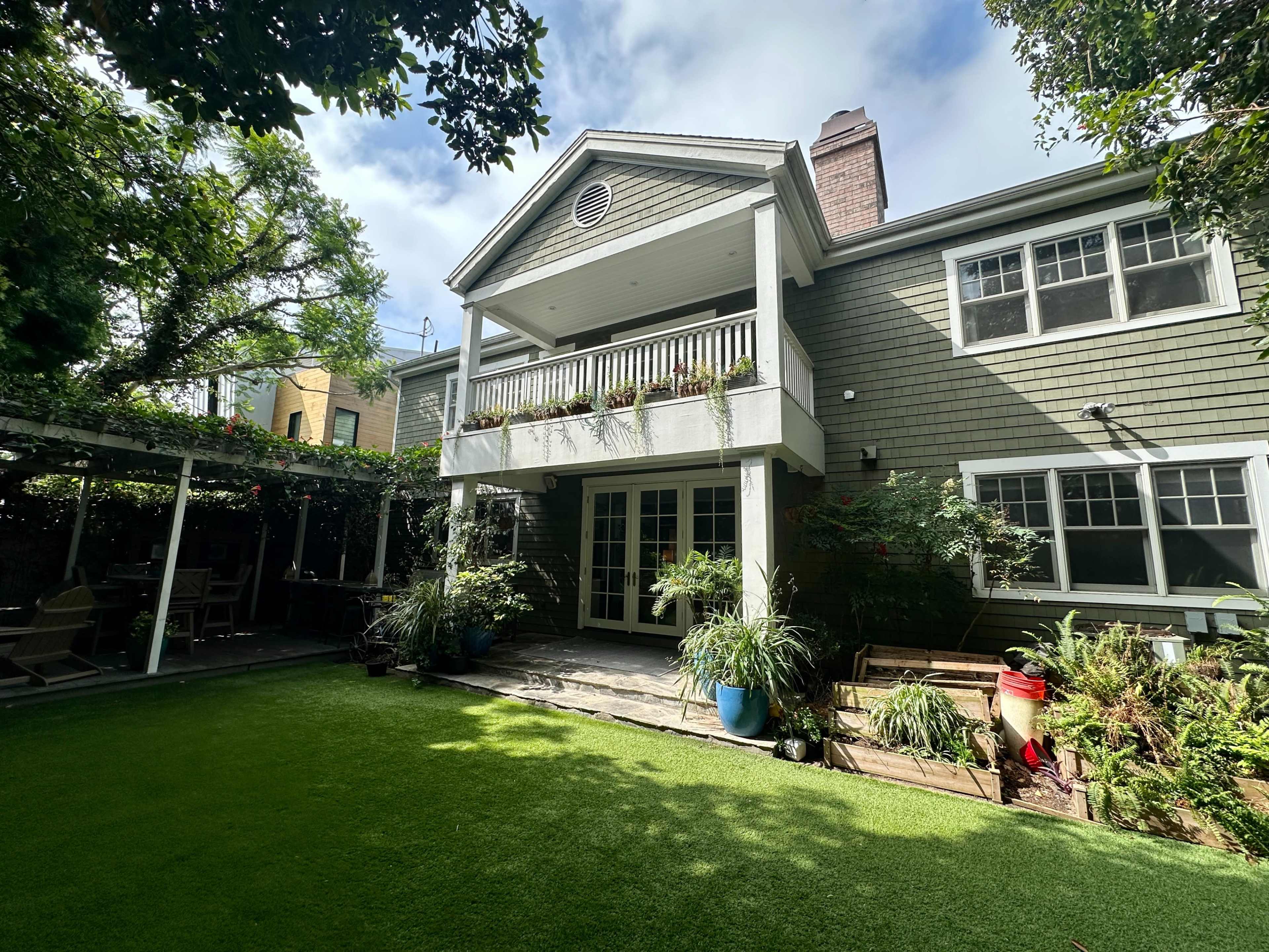 The image shows a two-story house with a covered balcony, surrounded by greenery and a well-maintained lawn.