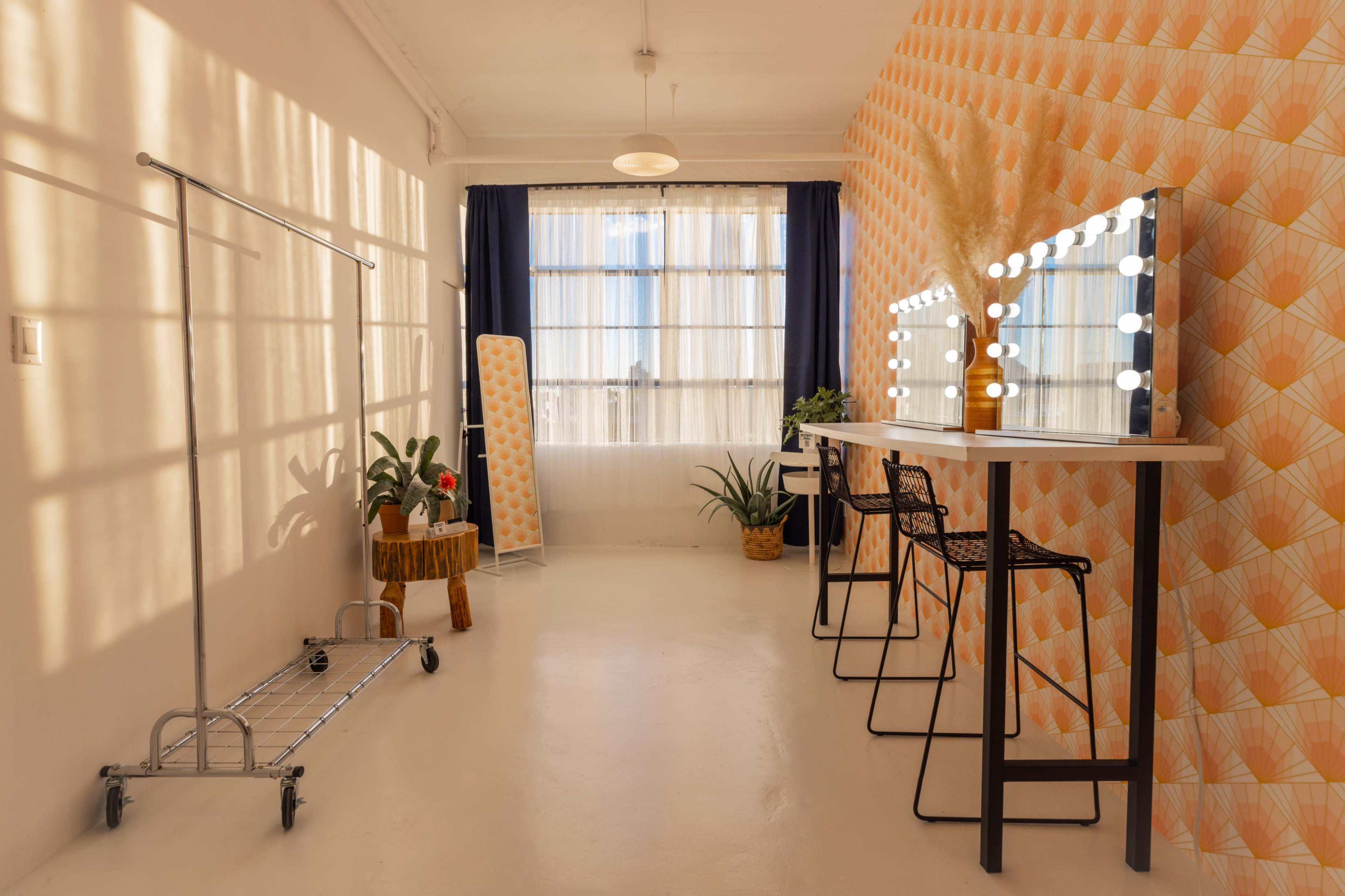 A brightly lit room with a makeup station featuring large illuminated mirrors, a clothing rack, and several plants against an orange patterned wall.