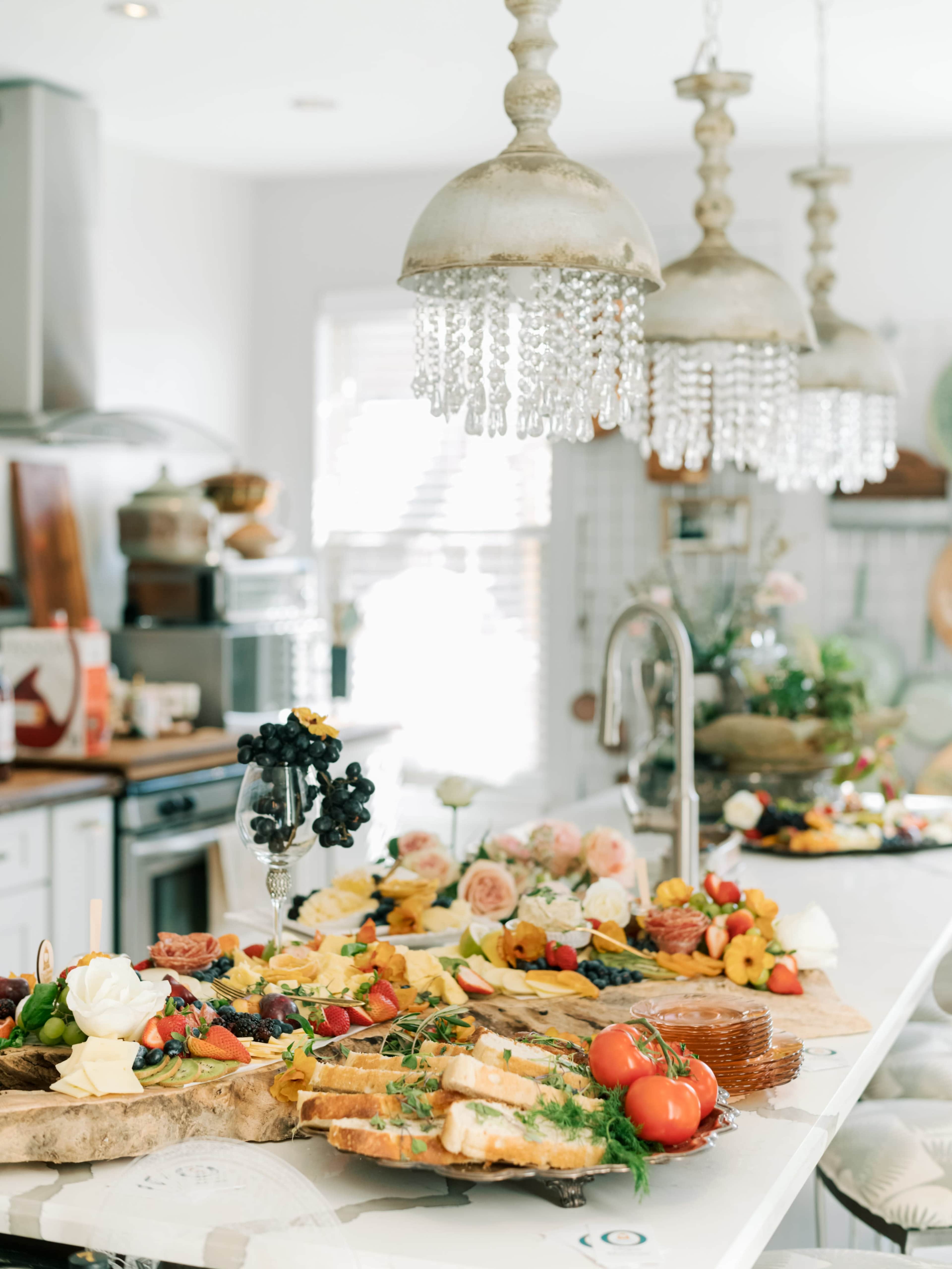 A large assortment of colorful food items, including fruits, cheeses, and flowers, is arranged on a marble countertop beneath decorative pendant lights.