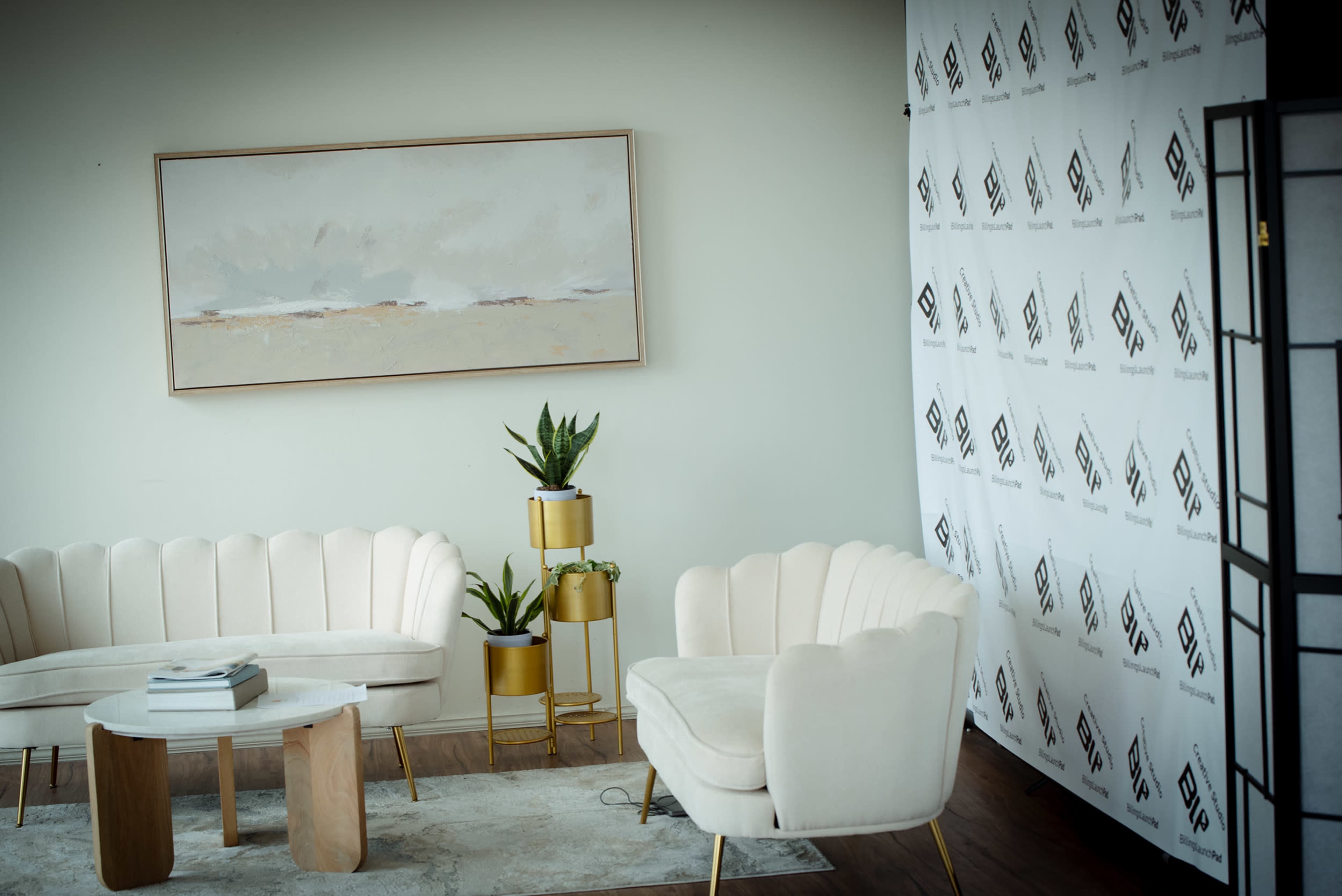 The image shows a modern indoor seating area featuring cream-colored sofas, a round coffee table, and potted plants against a light wall adorned with a patterned backdrop.