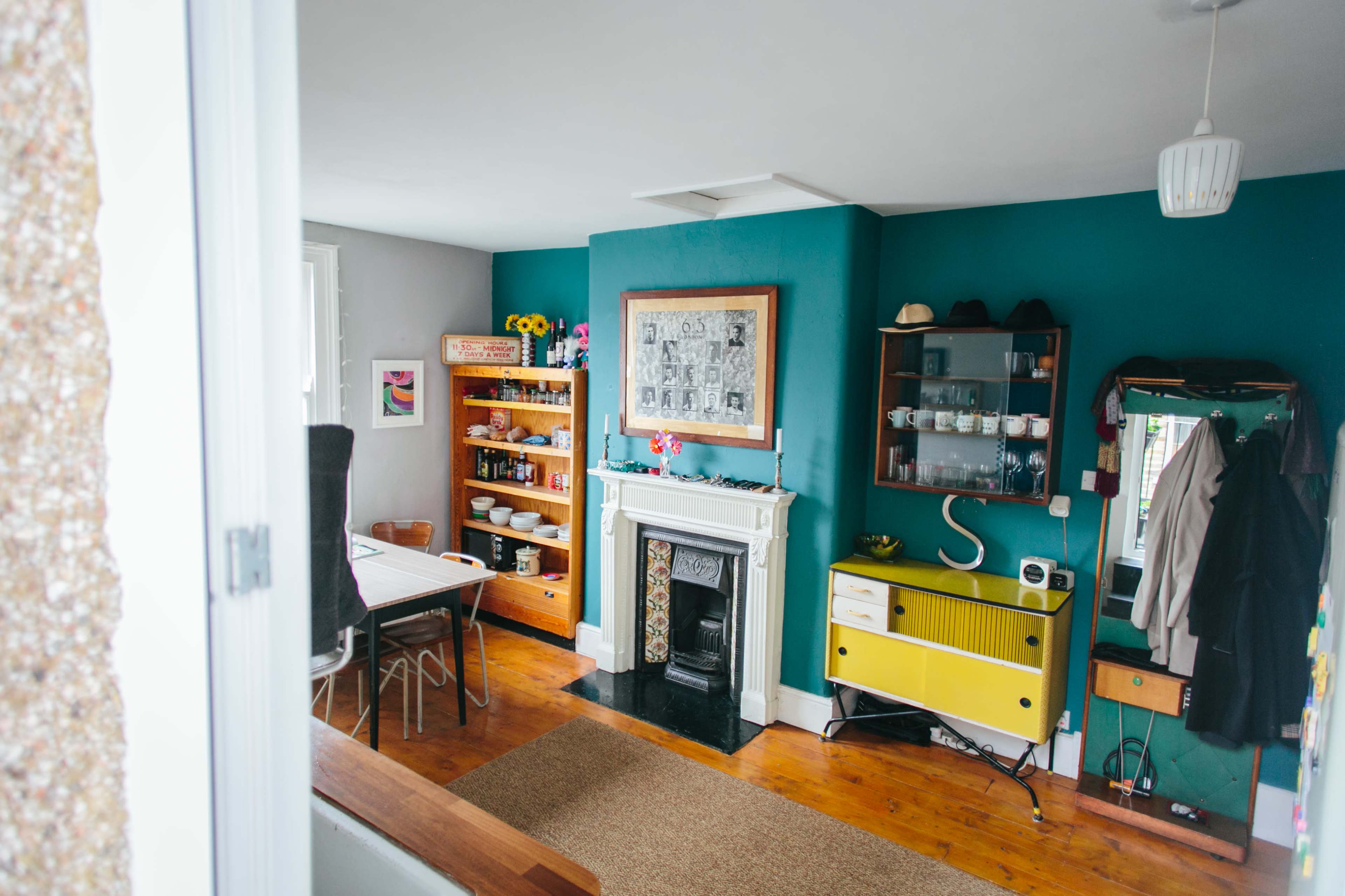The image shows a bright living space with a teal wall, a wooden floor, and furniture including a yellow sideboard and a dining table.