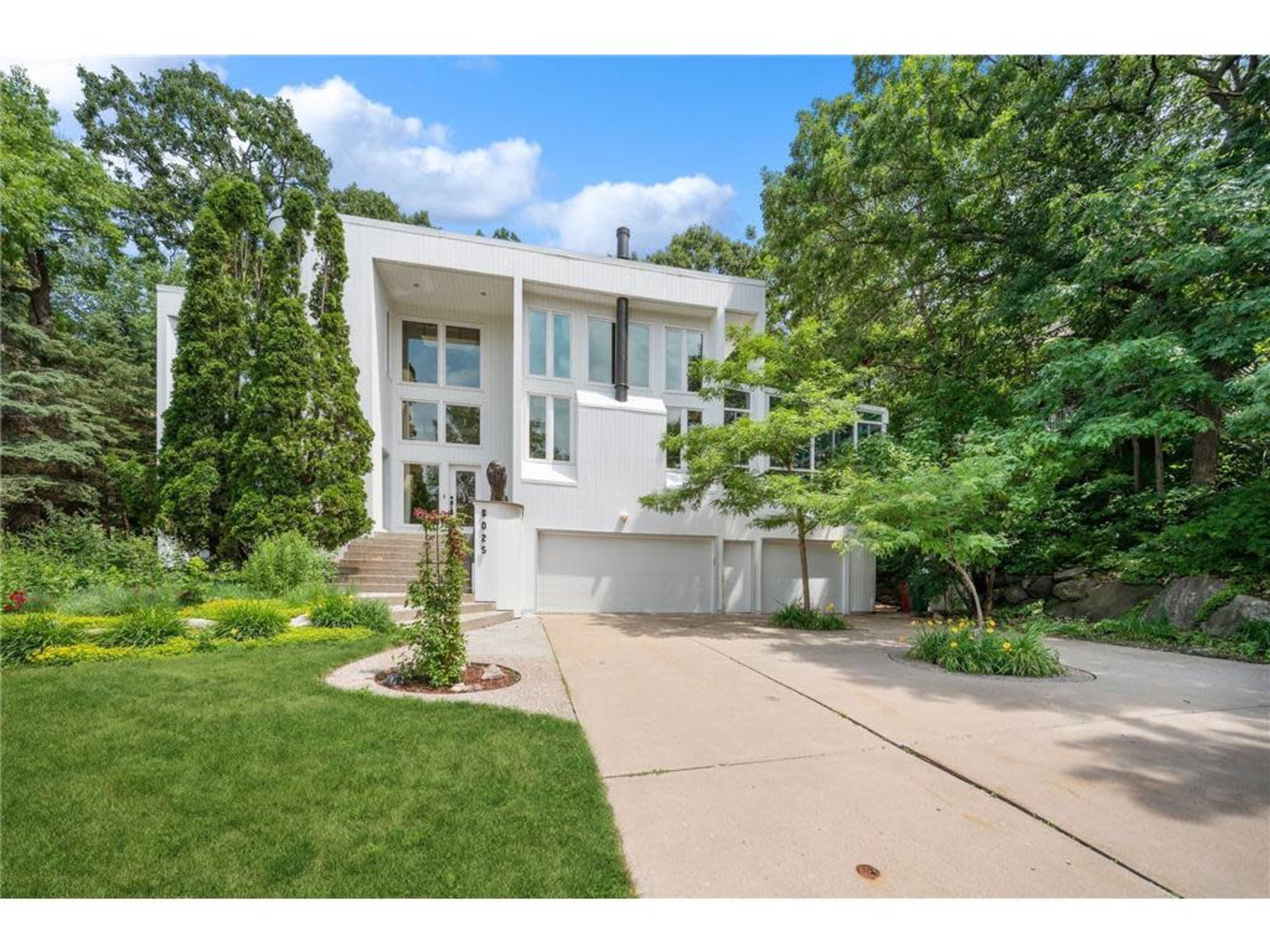 A modern two-story house with large windows sits on a landscaped lot featuring green trees and a well-maintained lawn.