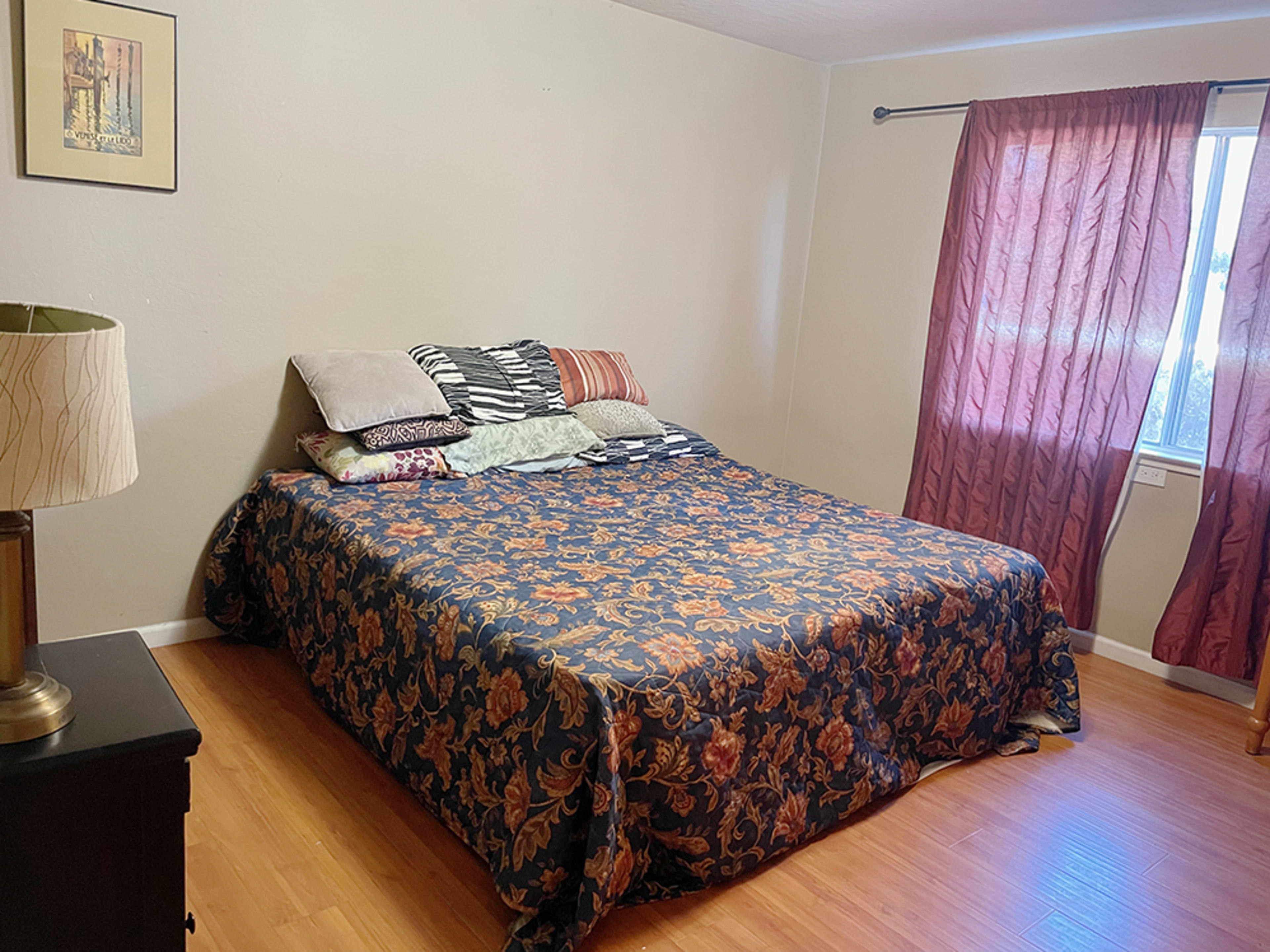A bedroom with a bed covered in a floral patterned blanket, several pillows atop it, and a window with red curtains.