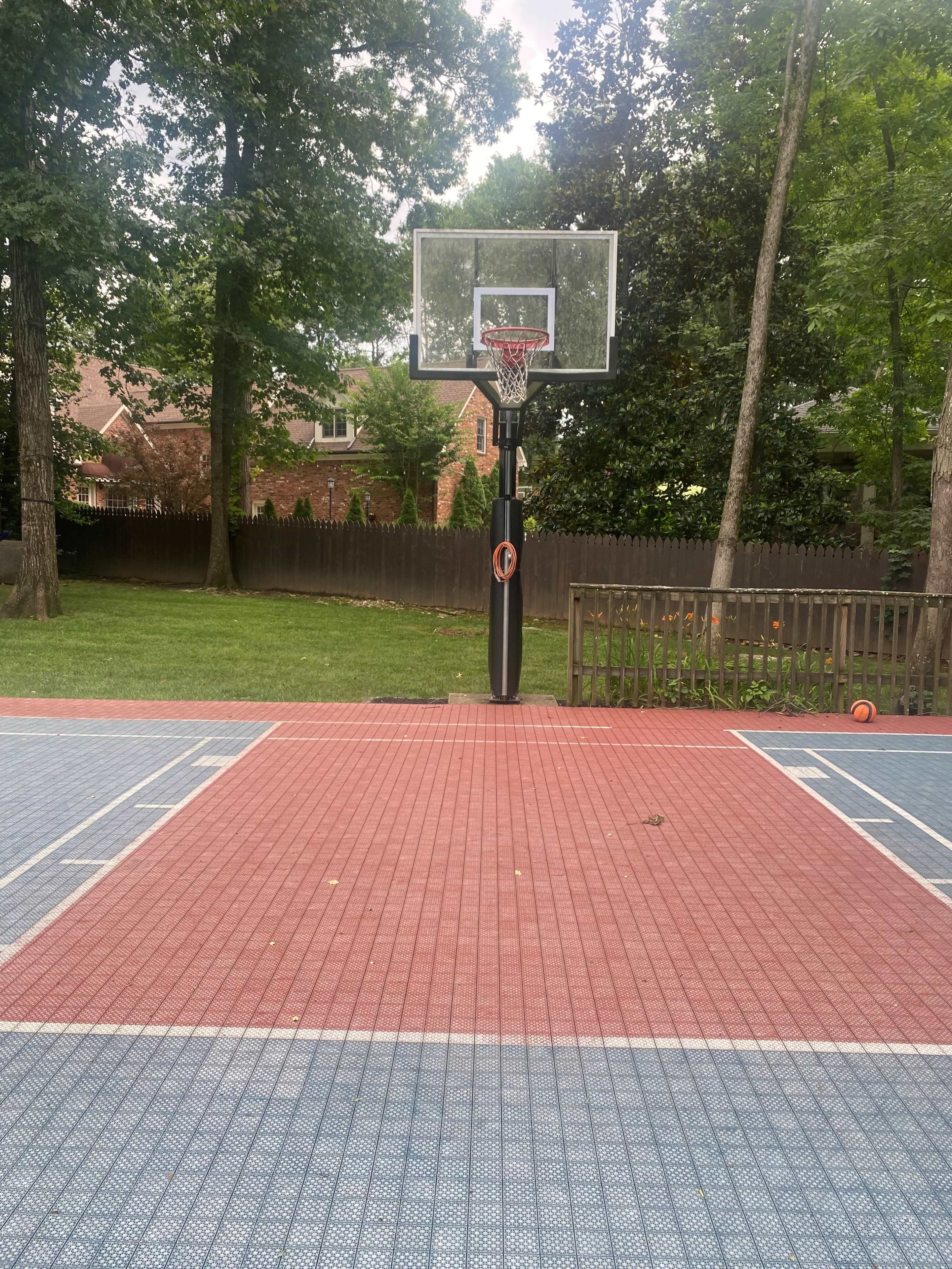 A basketball court with a hoop surrounded by trees and a wooden fence in the background.