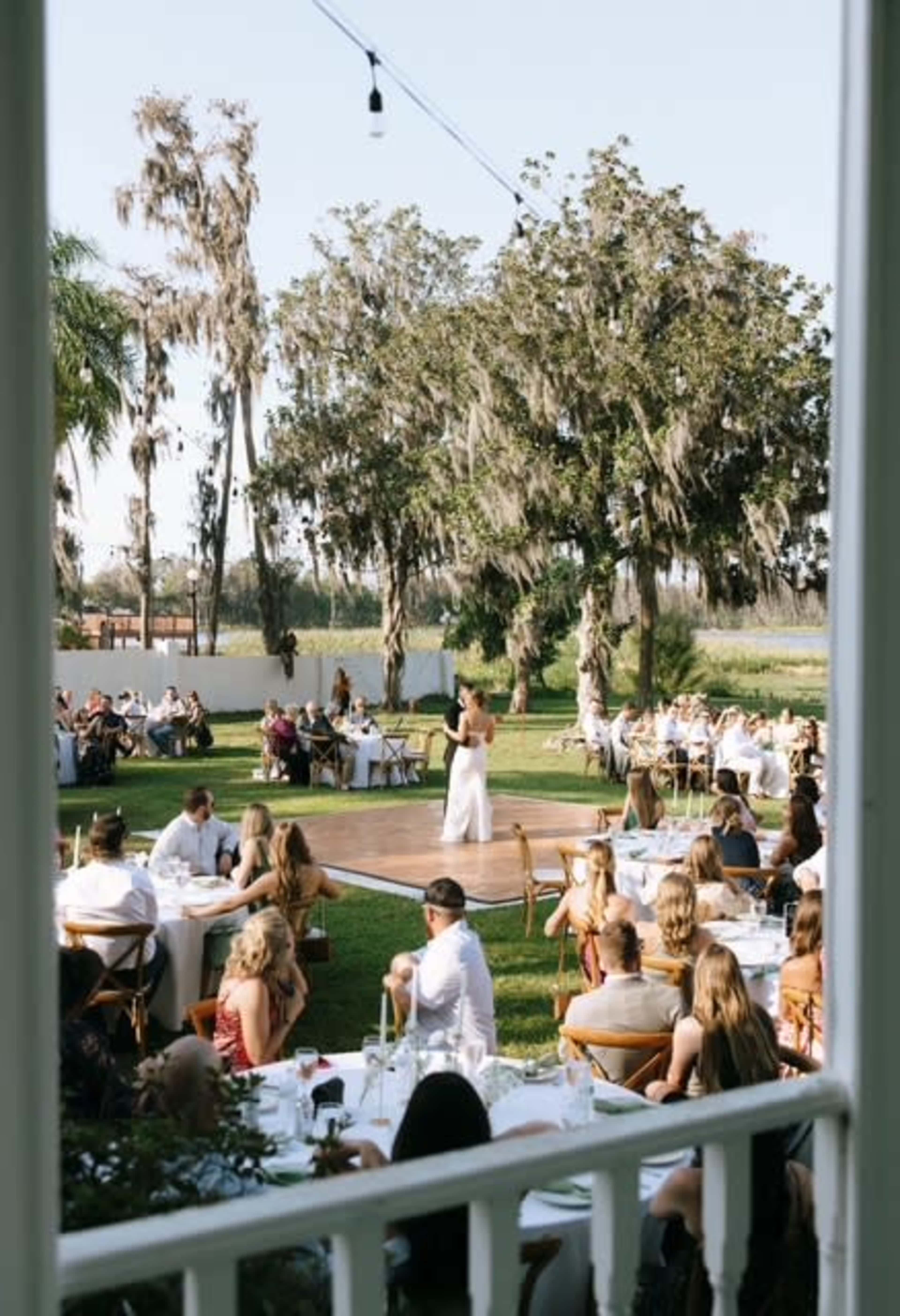 A couple dances on a wooden stage while guests watch and dine at an outdoor event surrounded by trees.