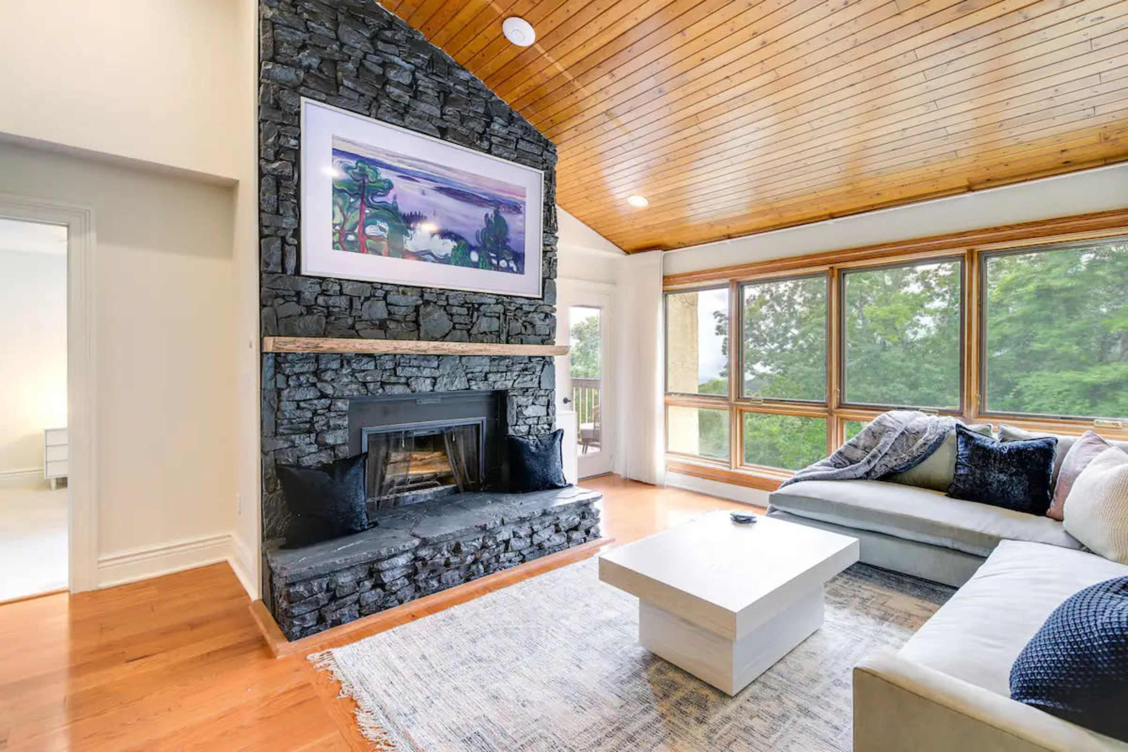 A living room with a stone fireplace, a sloped wooden ceiling, large windows, and a light-colored sofa facing a coffee table.