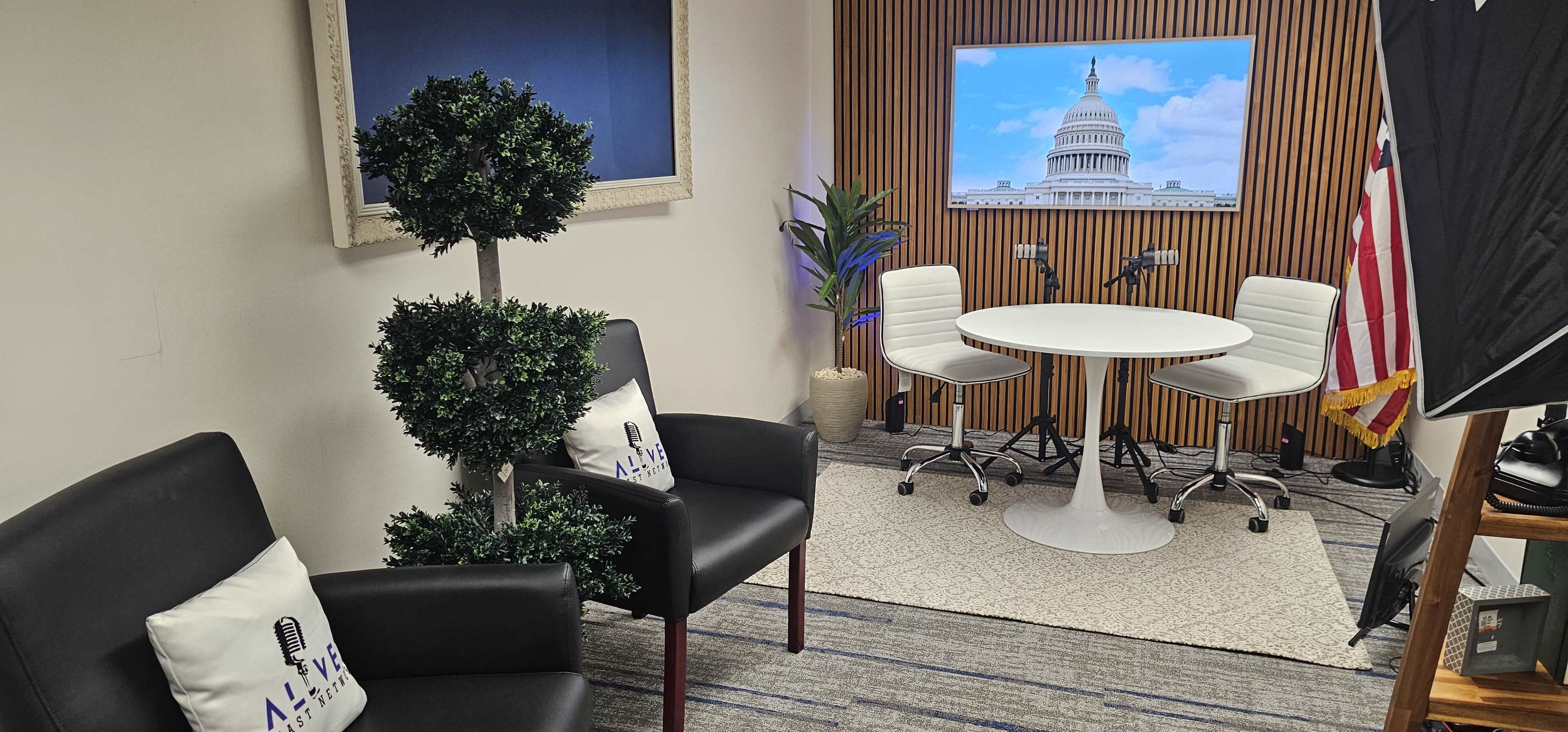 The image shows a modern studio setup with two white chairs and a round table in front of a screen displaying the U.S. Capitol, alongside artificial plants and a flag.