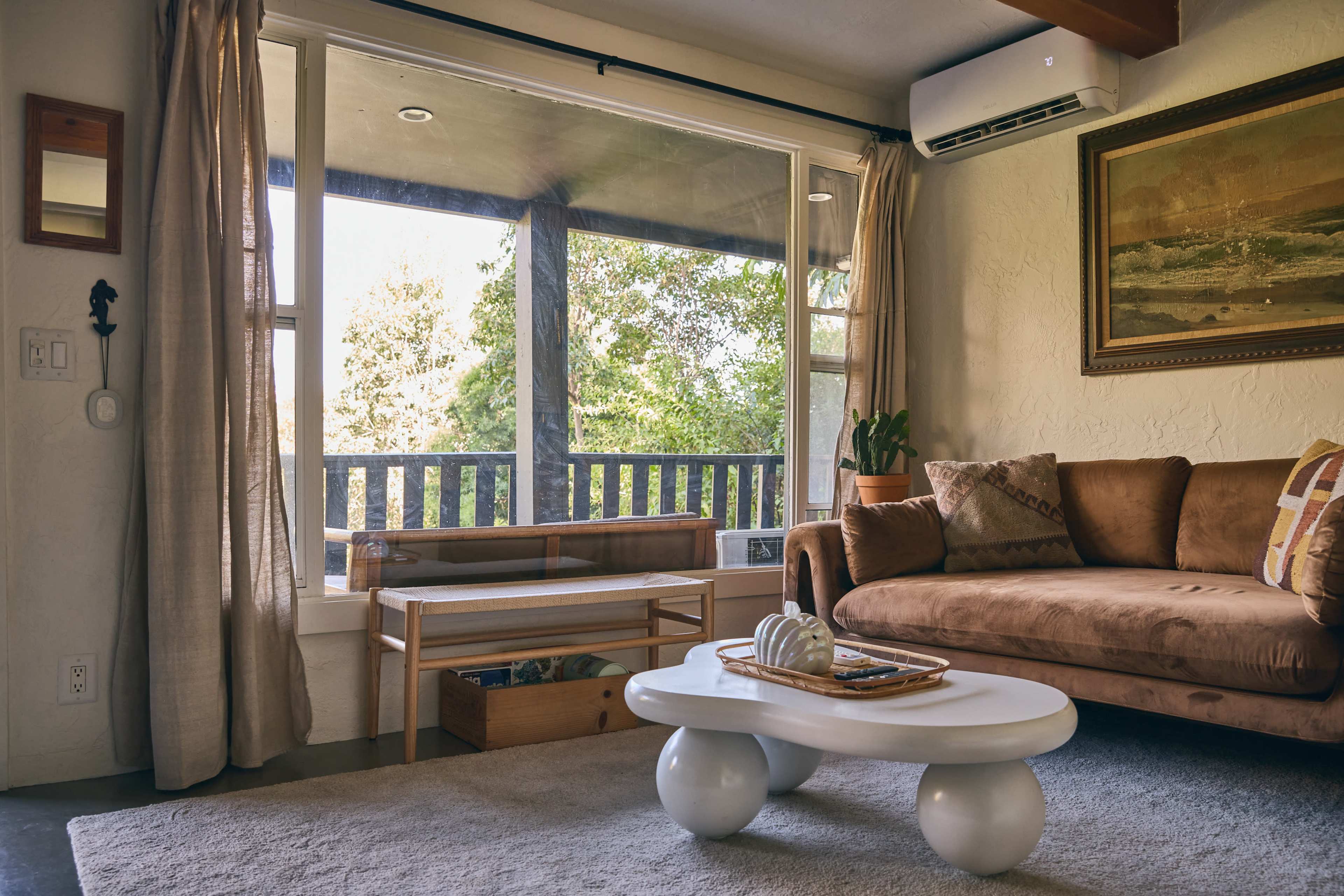 A cozy living room features a brown sofa, a round white coffee table, and large windows that provide a view of the outdoors.